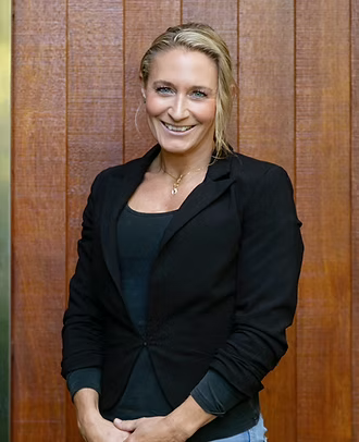 Portrait of a woman with blonde hair, smiling, wearing a black blazer over a dark top, standing in front of wooden paneling.
