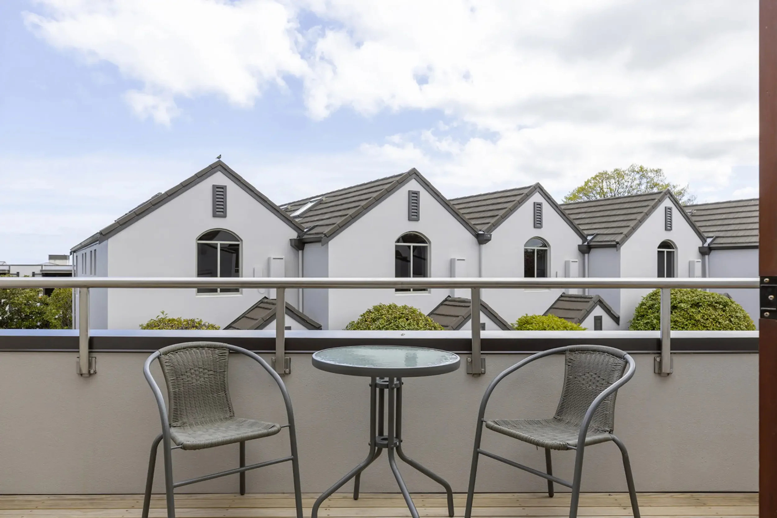 Balcony with two outdoor chairs and a glass table, overlooking white houses with gray roofs and green trees under a partly cloudy sky.