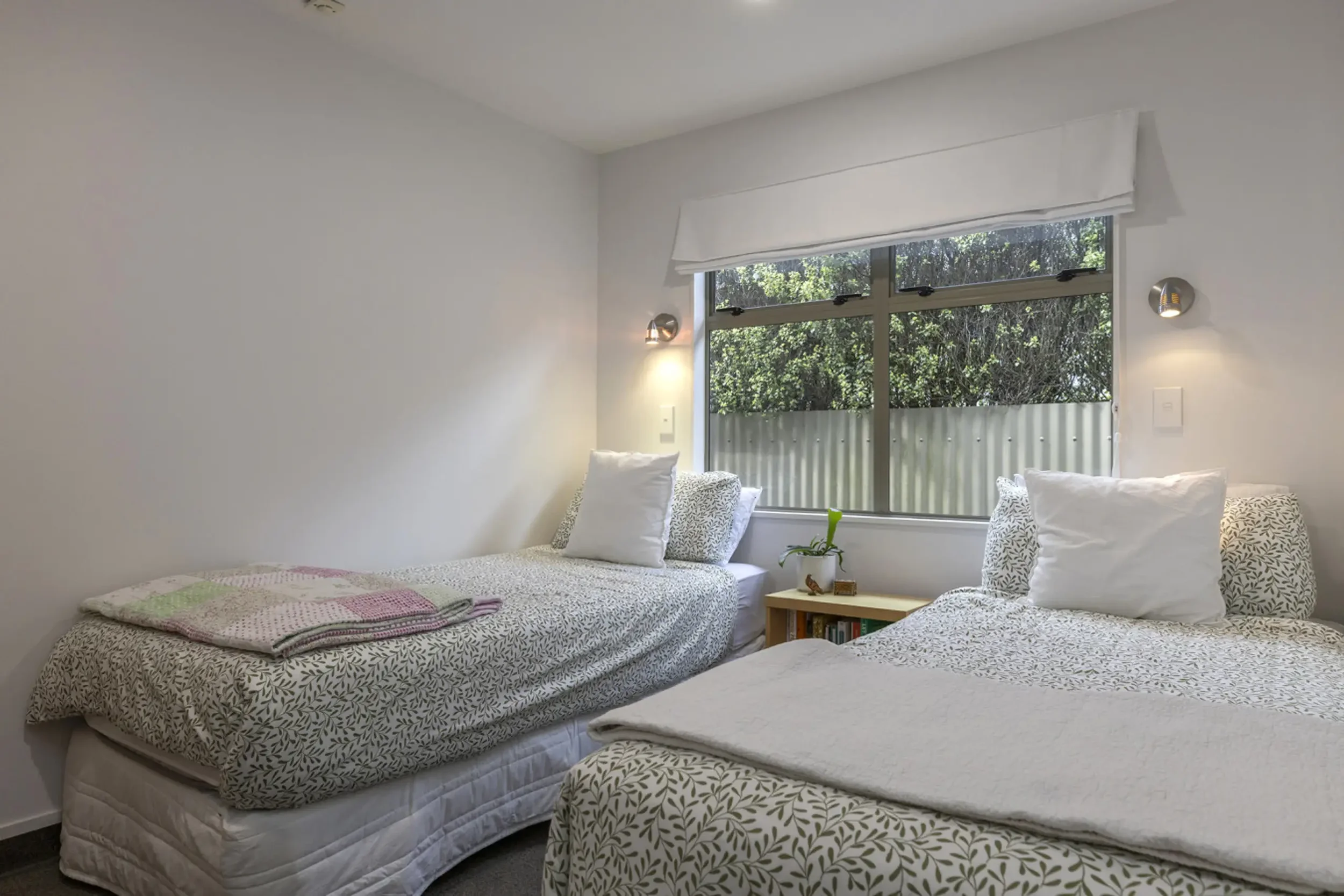 A bedroom with two single beds, white bedding with leaf patterns, stuffed pillows, a nightstand with a potted plant, and a window showing outdoor greenery.