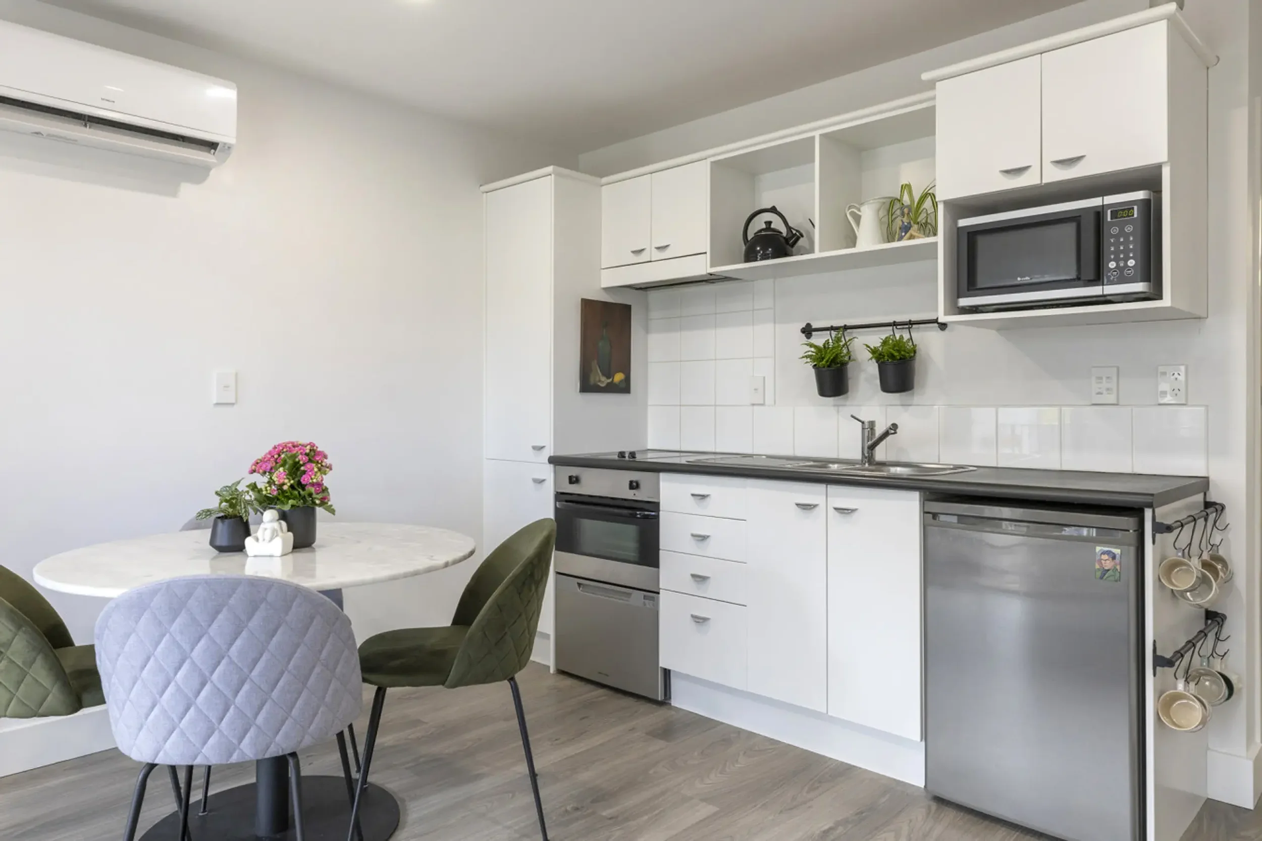 Kitchen with white cabinets, black teapot, microwave, small refrigerator, plants, dining table with chairs, air conditioner on wall.