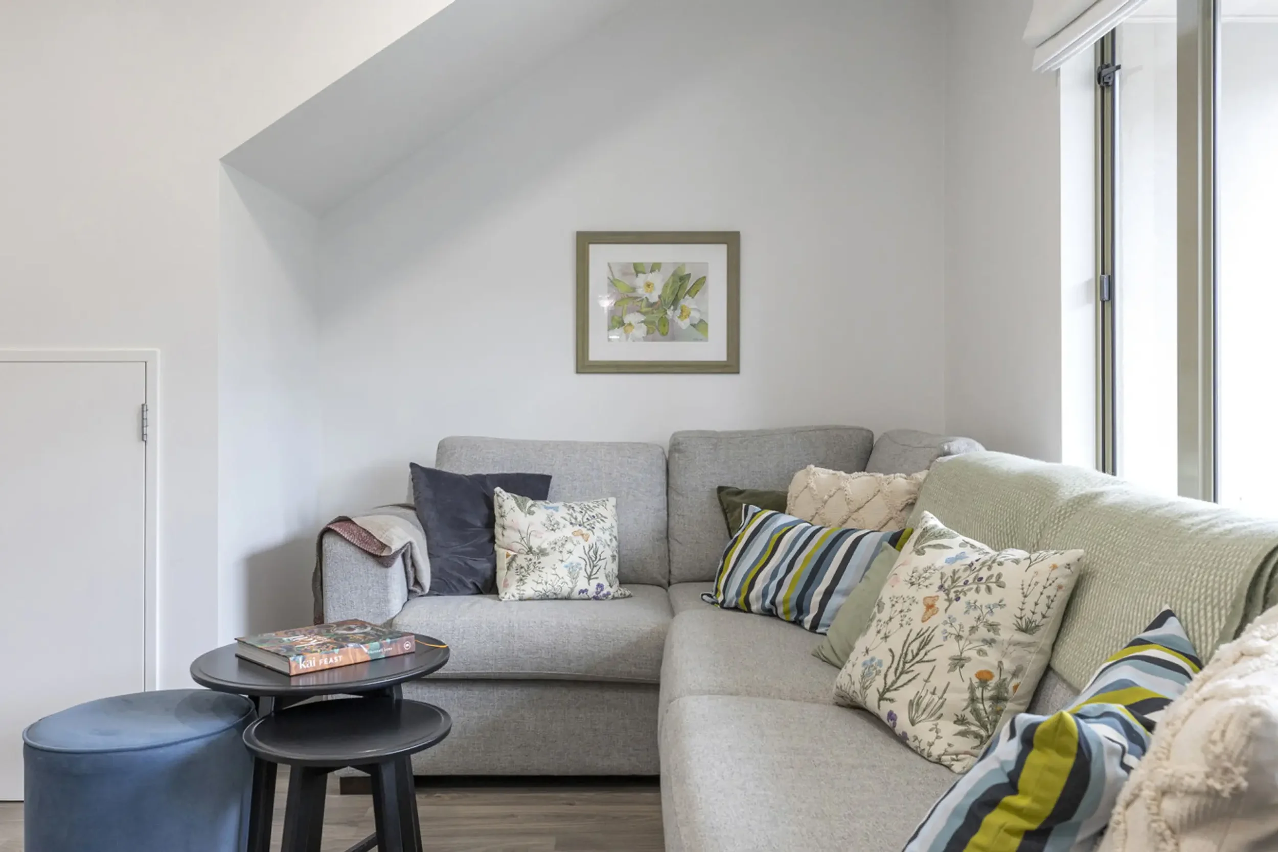 Living room corner with light gray sectional sofa decorated with patterned pillows, a small black side table with books, a round blue ottoman, and framed artwork of flowers on white wall next to large window.