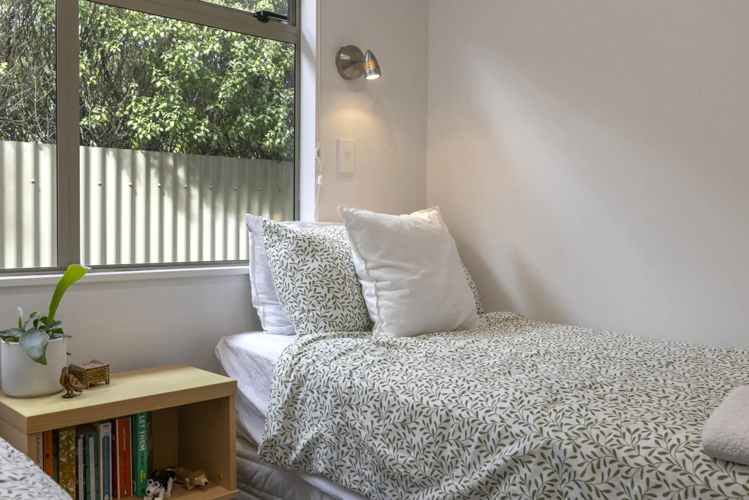 A neatly made bed with white sheets and patterned pillowcases, located near a window with trees outside, in a brightly lit bedroom.