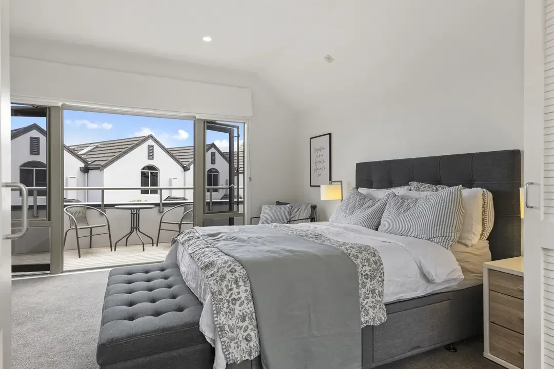 A bedroom with a large bed, pillows, and a gray headboard, next to a nightstand with a lamp, and a balcony with outdoor seating.