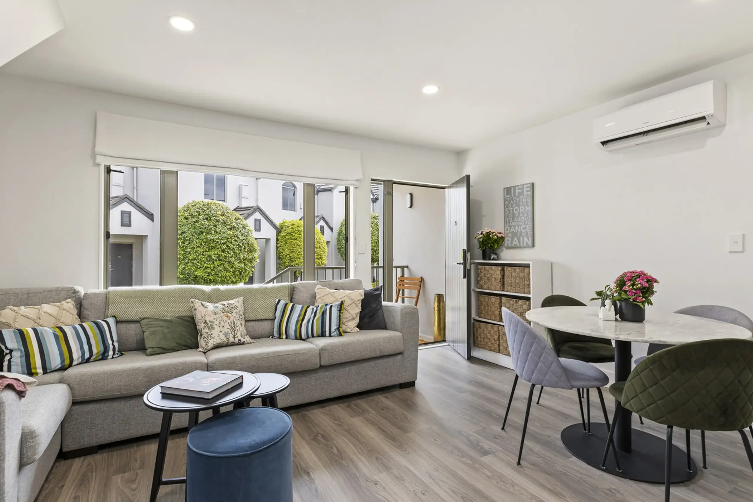 Bright, modern living room with gray sectional sofa, round dining table with chairs, and large window/door leading to a small outdoor area with shrubs.
