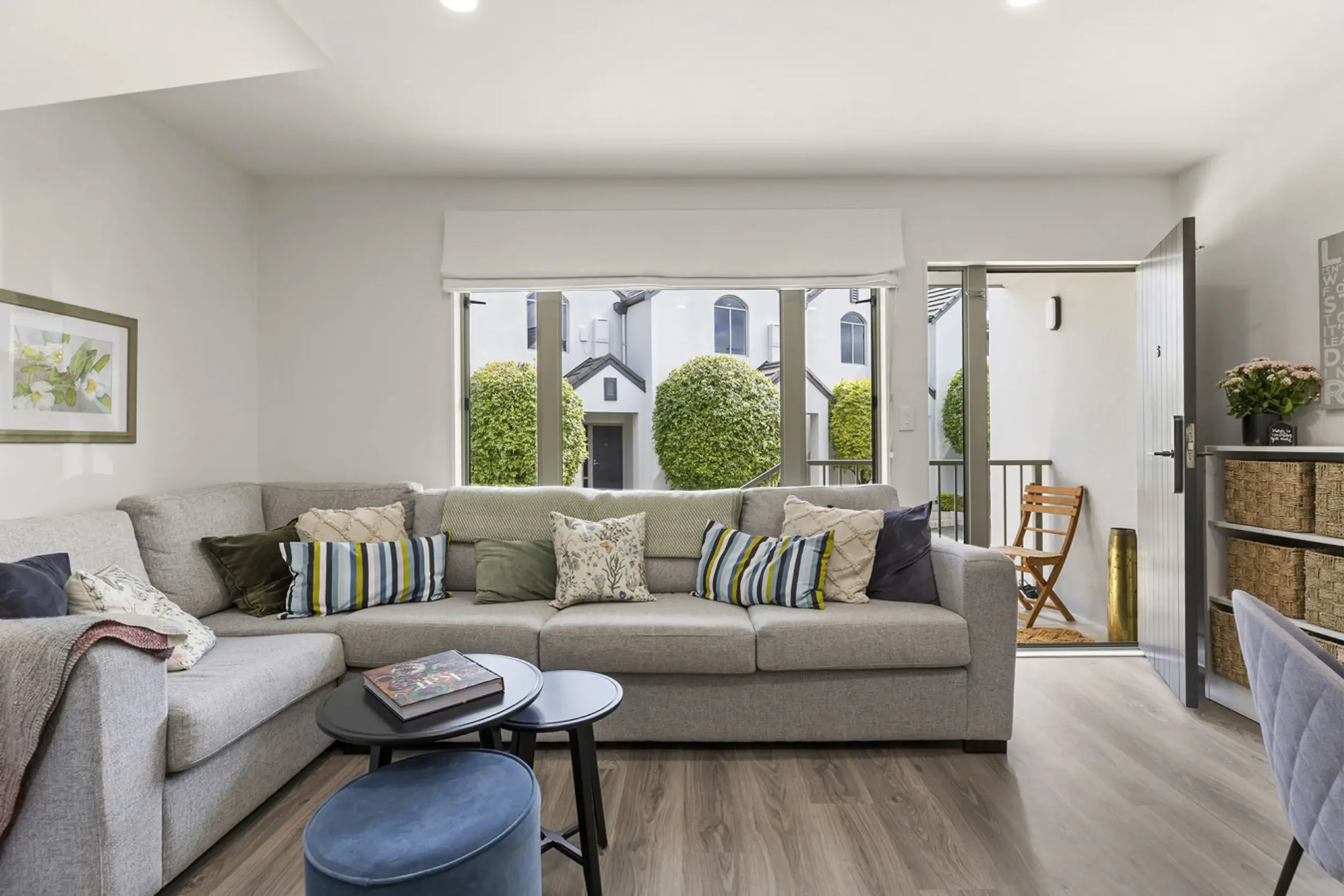 Living room with a beige couch adorned with multiple colorful throw pillows, a black coffee table with a magazine, and a large window with white blinds showing neatly trimmed green bushes outside.