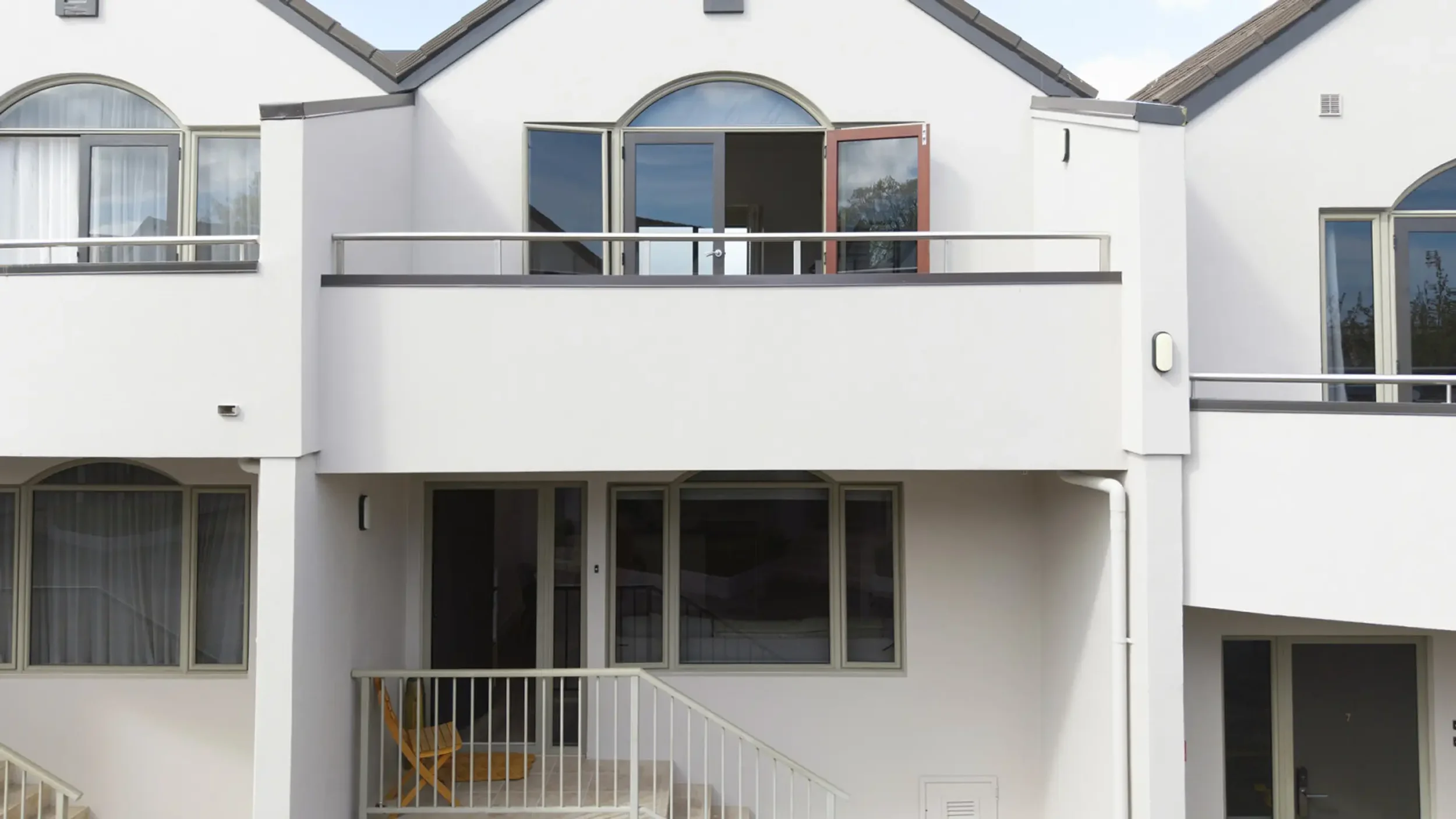 Front view of a modern white multi-story house with large windows, glass doors, and balconies. The house has a sloped roof and outdoor stairs leading to the entrance area.