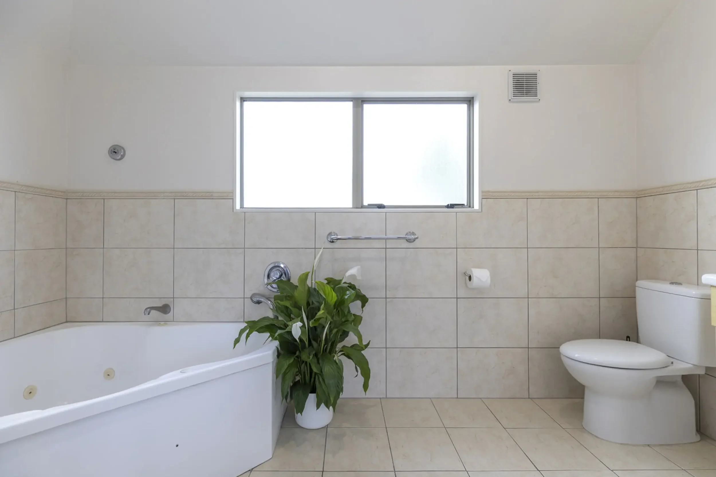 A bathroom with beige tiled walls and floor, a white bathtub with jets, a toilet, a window above the bathtub, a green potted plant next to the bathtub, and a toilet paper roll to the right of the plant.
