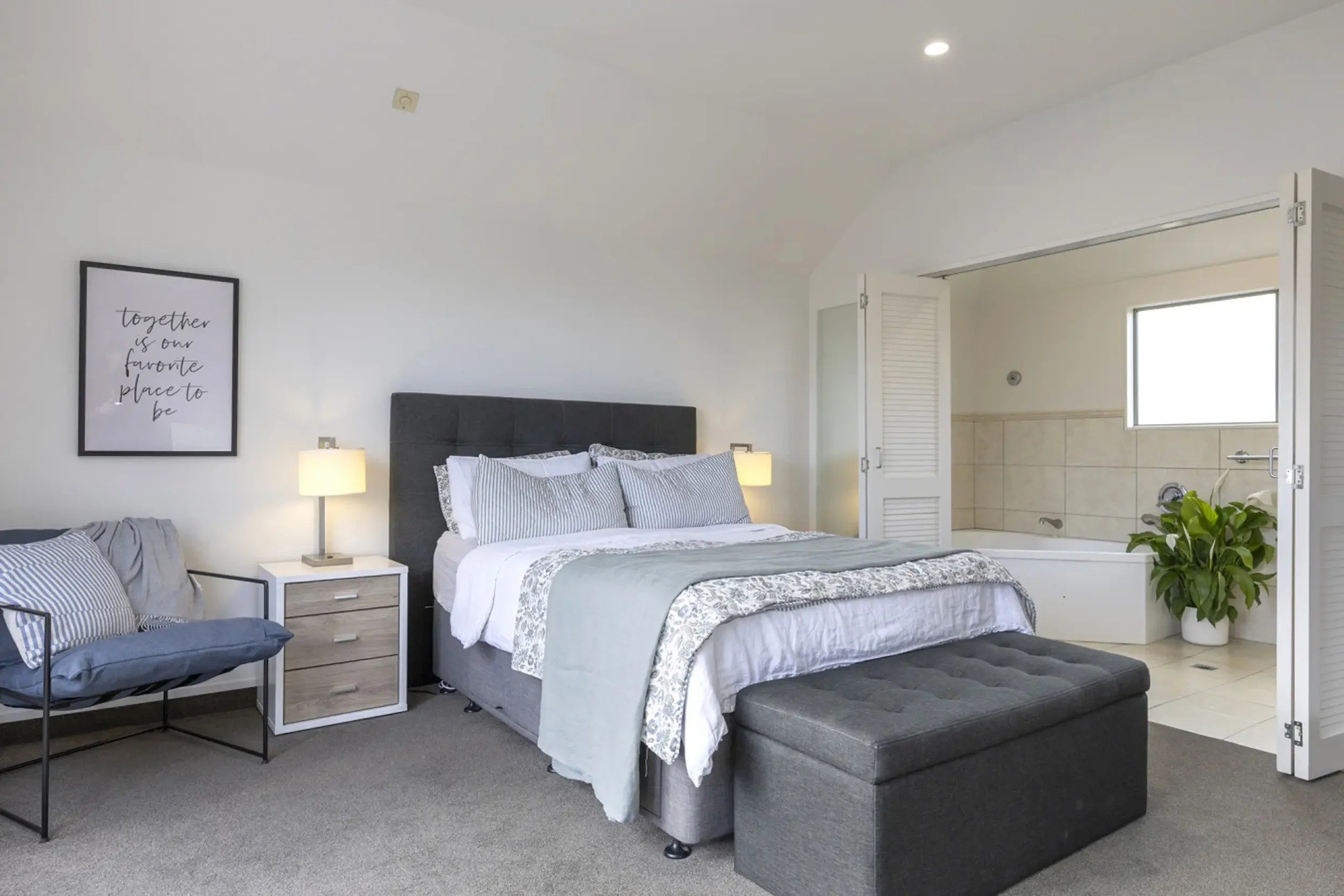 A bedroom with a bed, nightstand, and lamps, next to a bathroom with a bathtub, window, and potted plant.