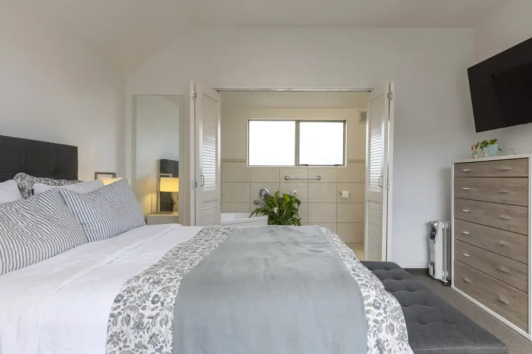 A bedroom with a bed, nightstand, dresser, and a window overlooking a bathroom with a plant and tiled walls.