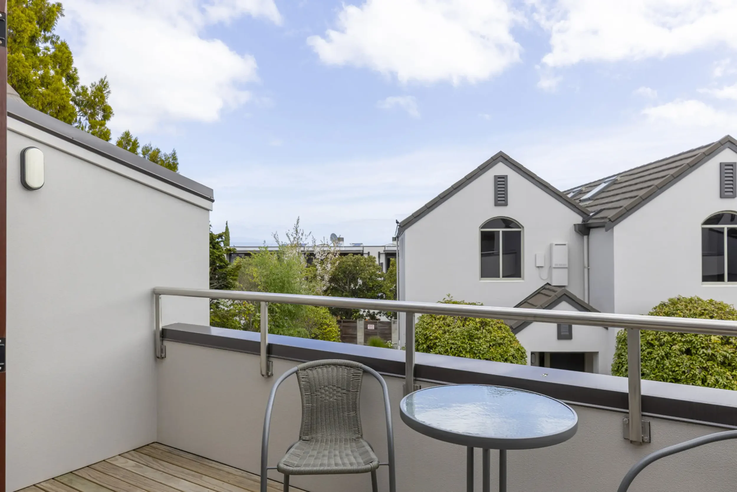 Balcony with a glass table and a chair overlooking a residential neighborhood with trees and houses under a partly cloudy sky.
