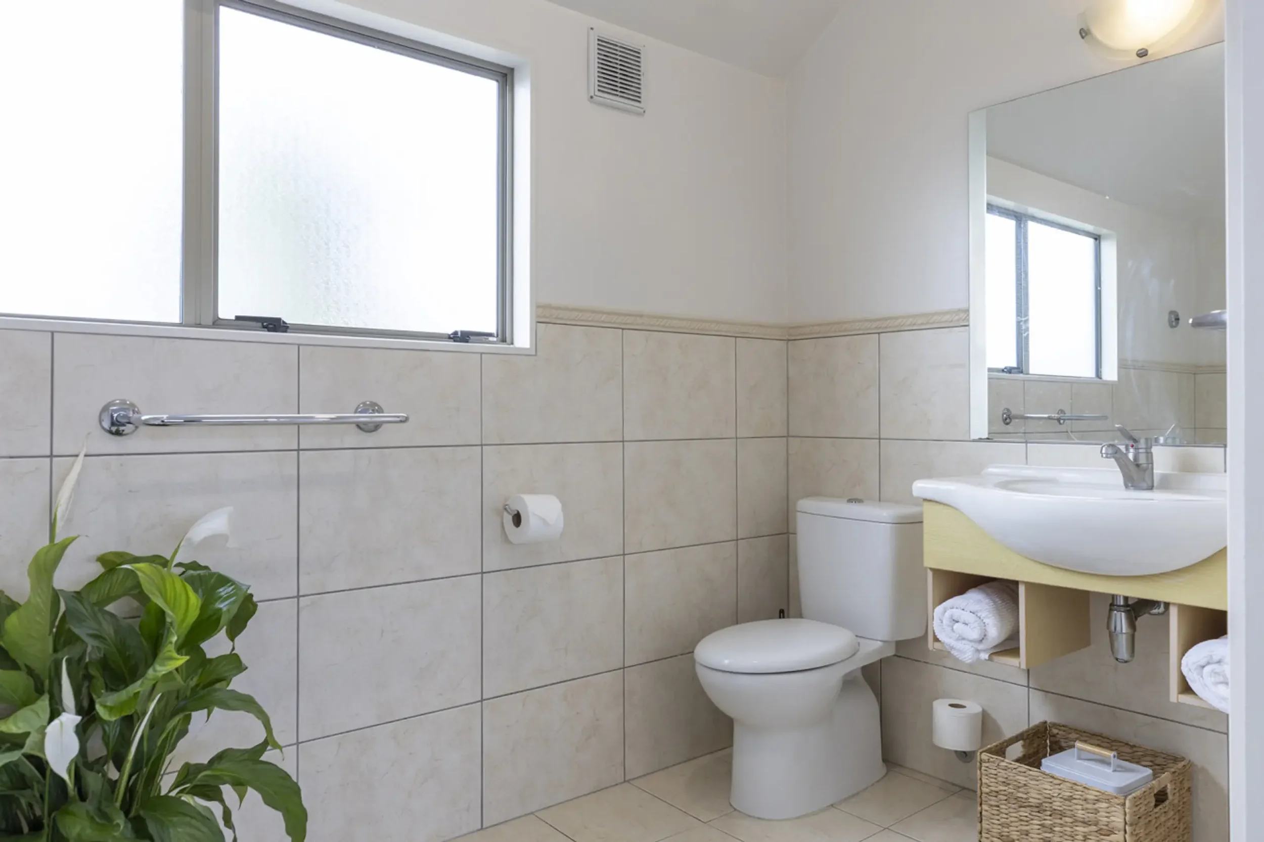 A clean bathroom with a white toilet, a small white sink with a mirror, a window with frosted glass, a plant in the corner, and a wicker basket on the floor.