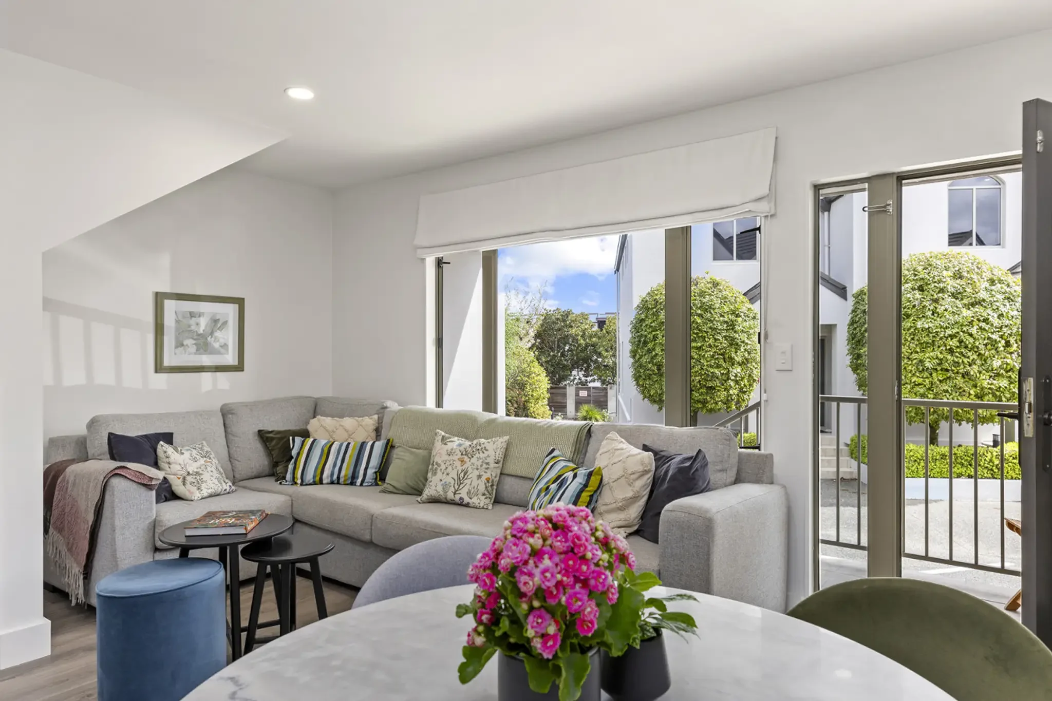 Bright living room with gray sectional sofa, multiple decorative pillows, a round marble table with pink flowers, and large windows with a view of green trees outside.