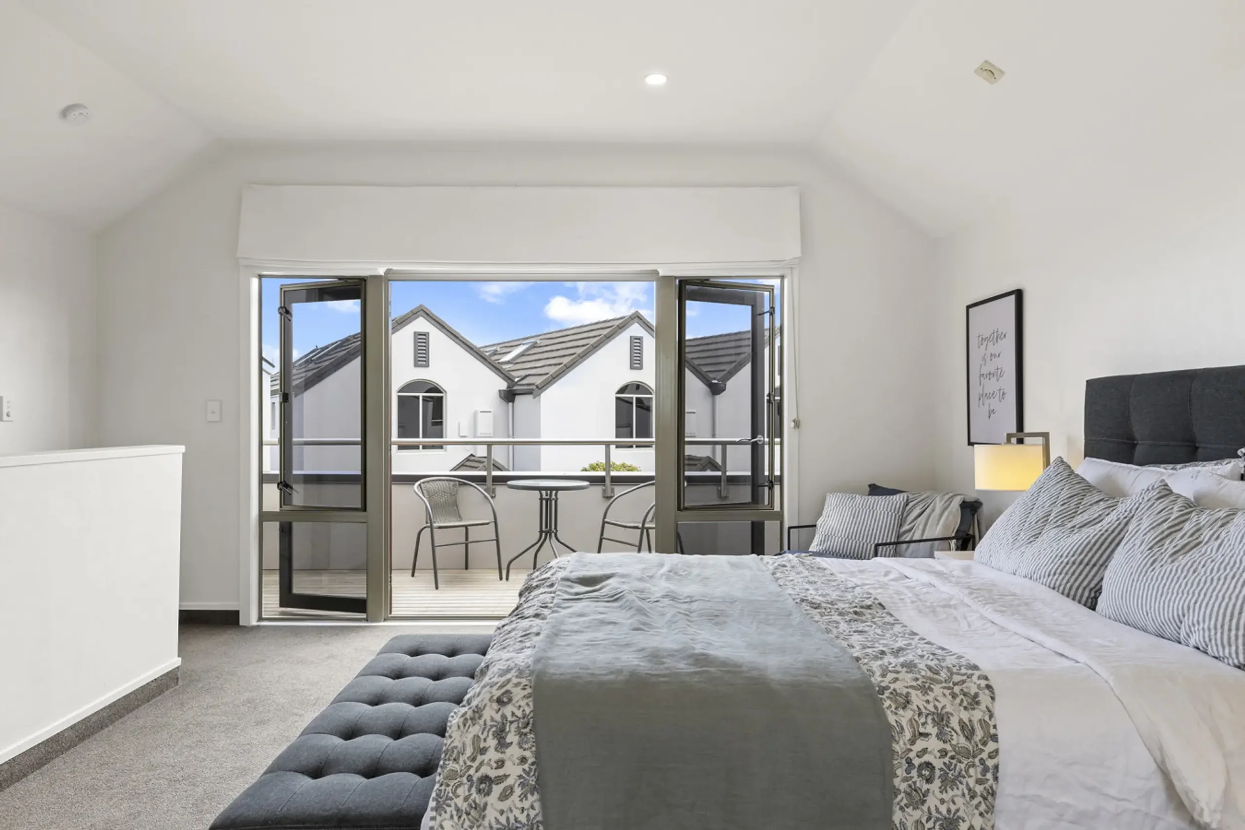 A bedroom with a bed, pillows, and a nightstand with a lamp. There is a balcony with two chairs and a small table outside, visible through open doors. The room has white walls and ceiling, and a gray carpet.