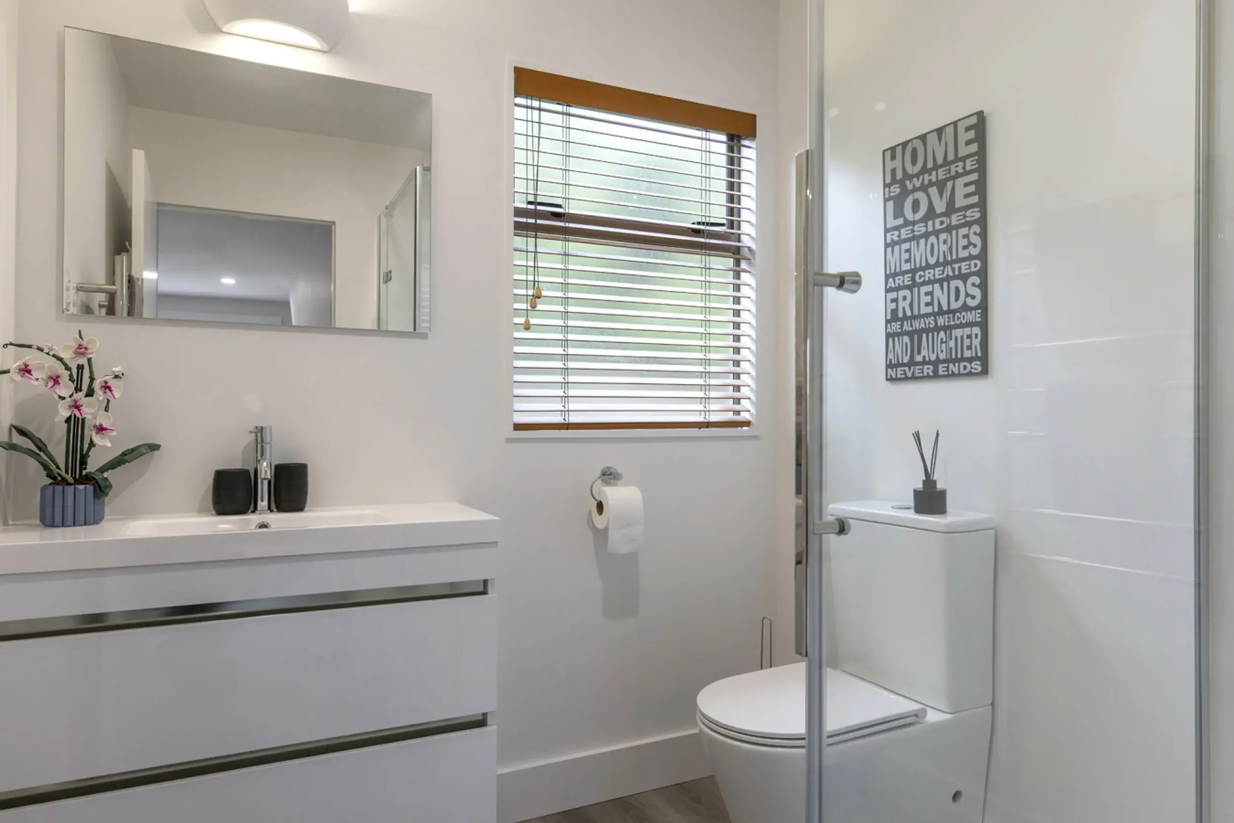 Modern bathroom with white vanity, mirror, orchid plant, black speakers, window with blinds, toilet, wall art, and shower area.