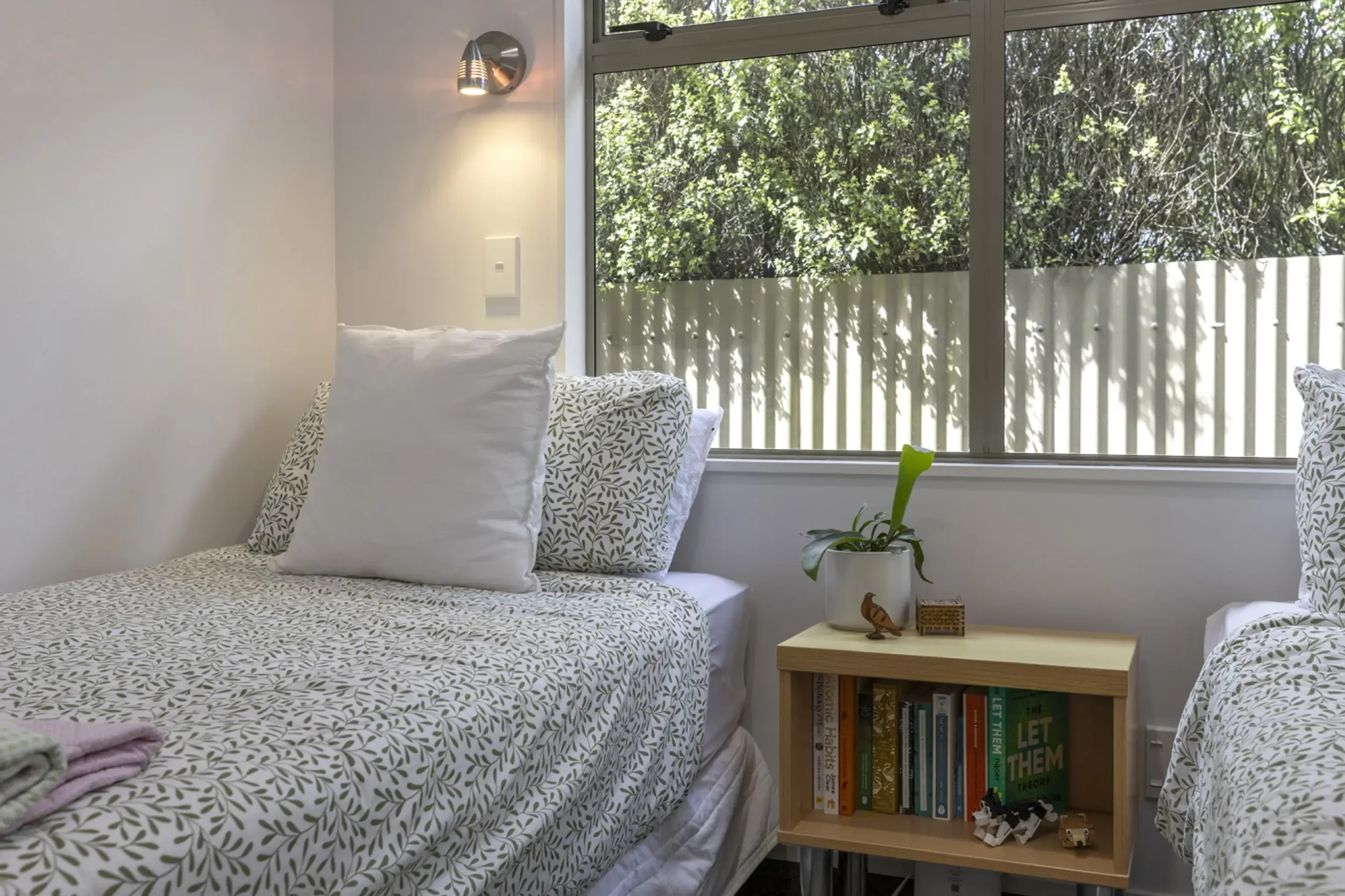 A bedroom with two beds featuring white and leafy patterned bedding and pillows, a small wooden bookshelf with books and decorative items, a white flowerpot with a green plant, and a large window with greenery outside.