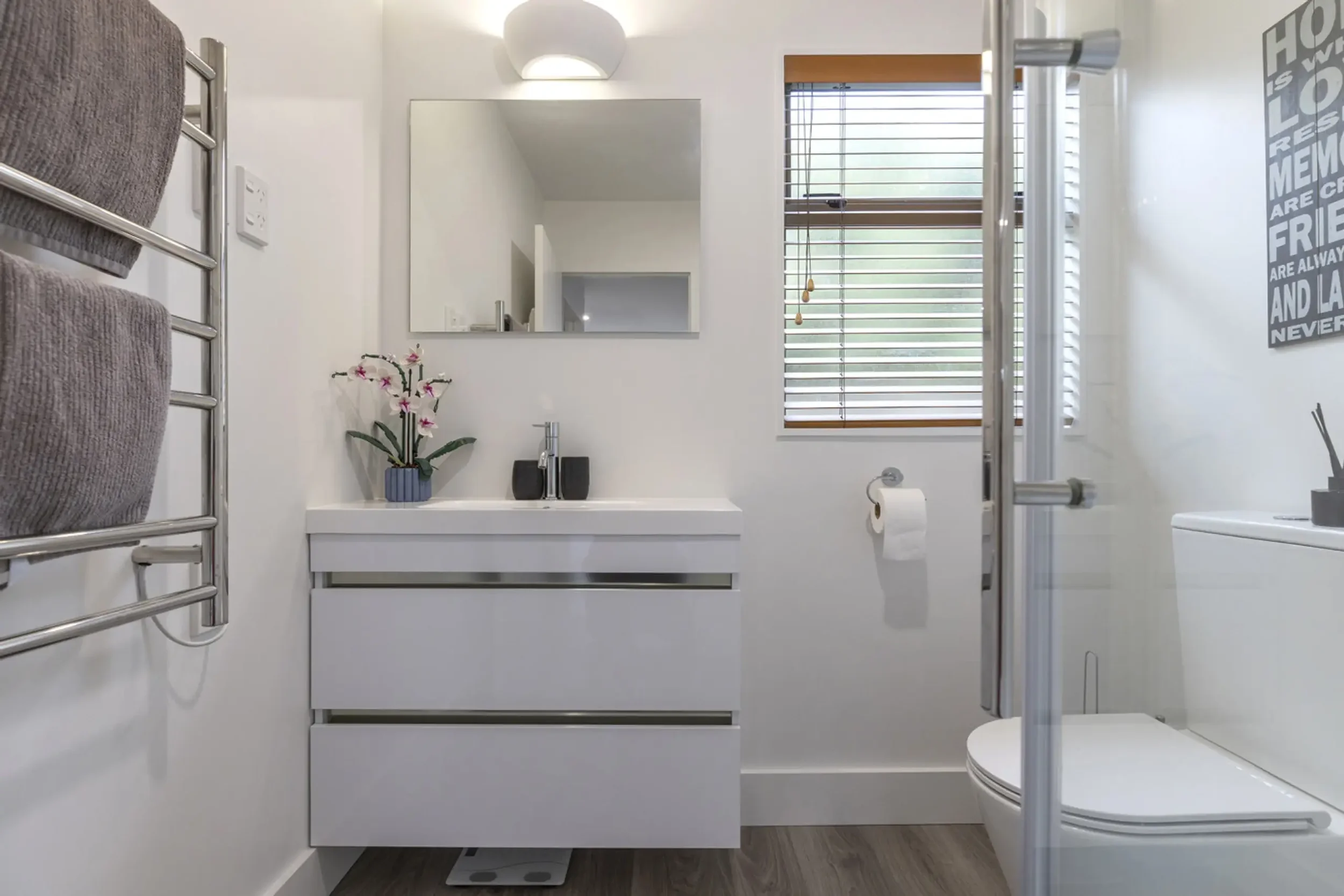 Minimalist bathroom with white walls, a small vanity with a mirror, a window with blinds, a toilet, a heated towel rail with towels, and a shower enclosure.
