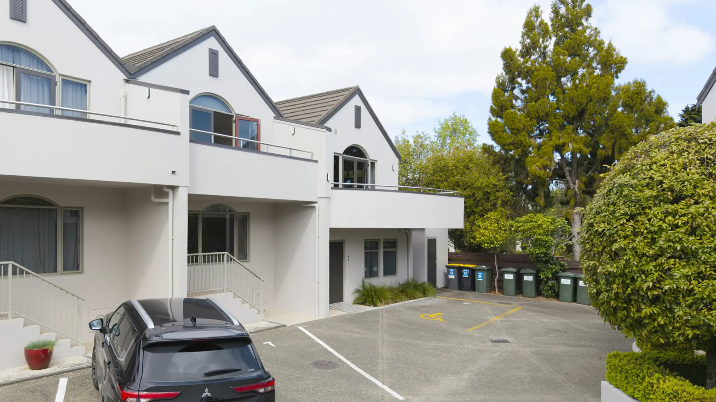 A residential apartment complex with white exterior walls and multiple balconies, parked car, and green trash bins, surrounded by trees.