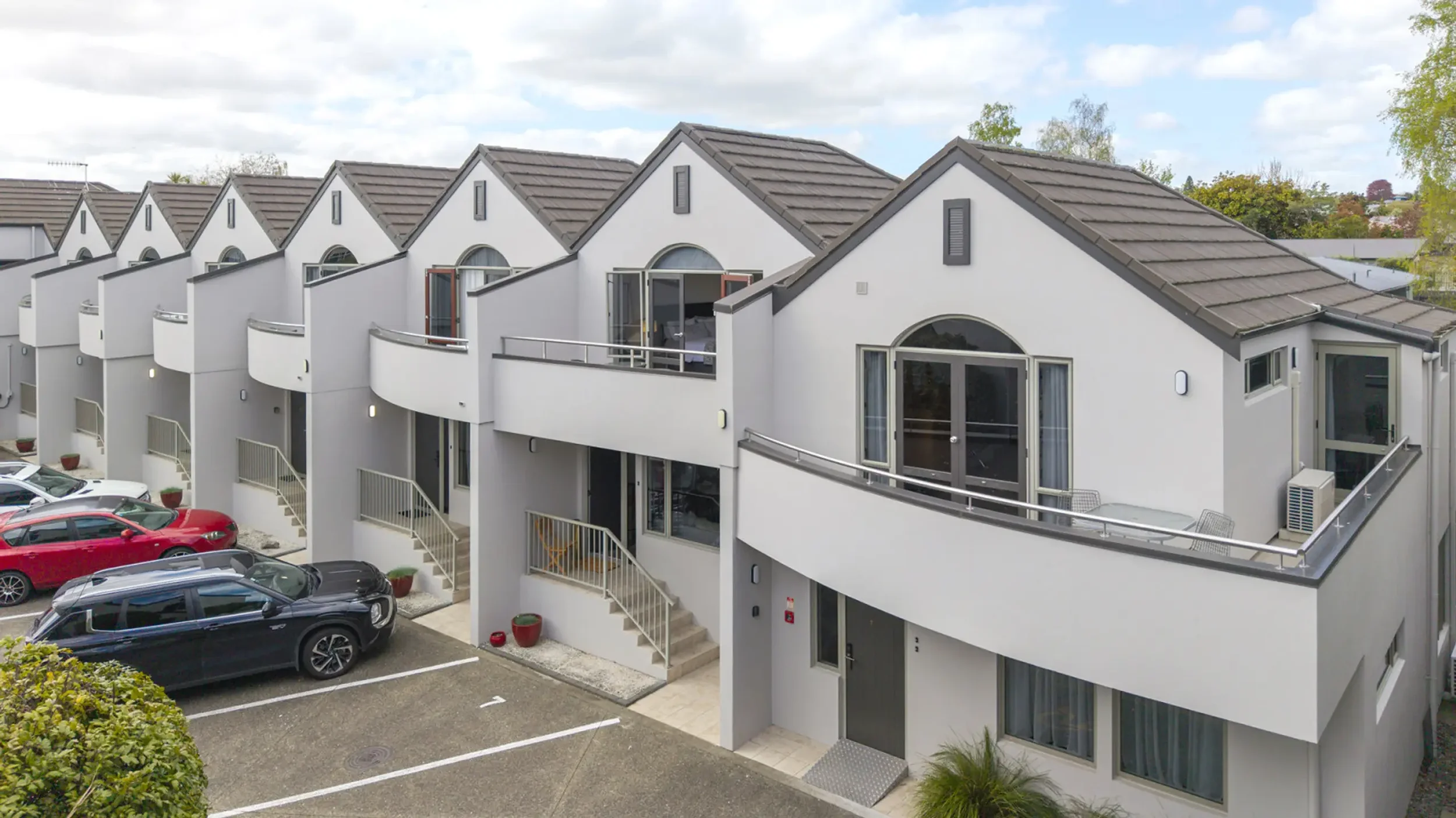 Row of modern white townhouses with grey tile roofs, small front stairs, and balconies, parked cars in the foreground, partly cloudy sky in the background.