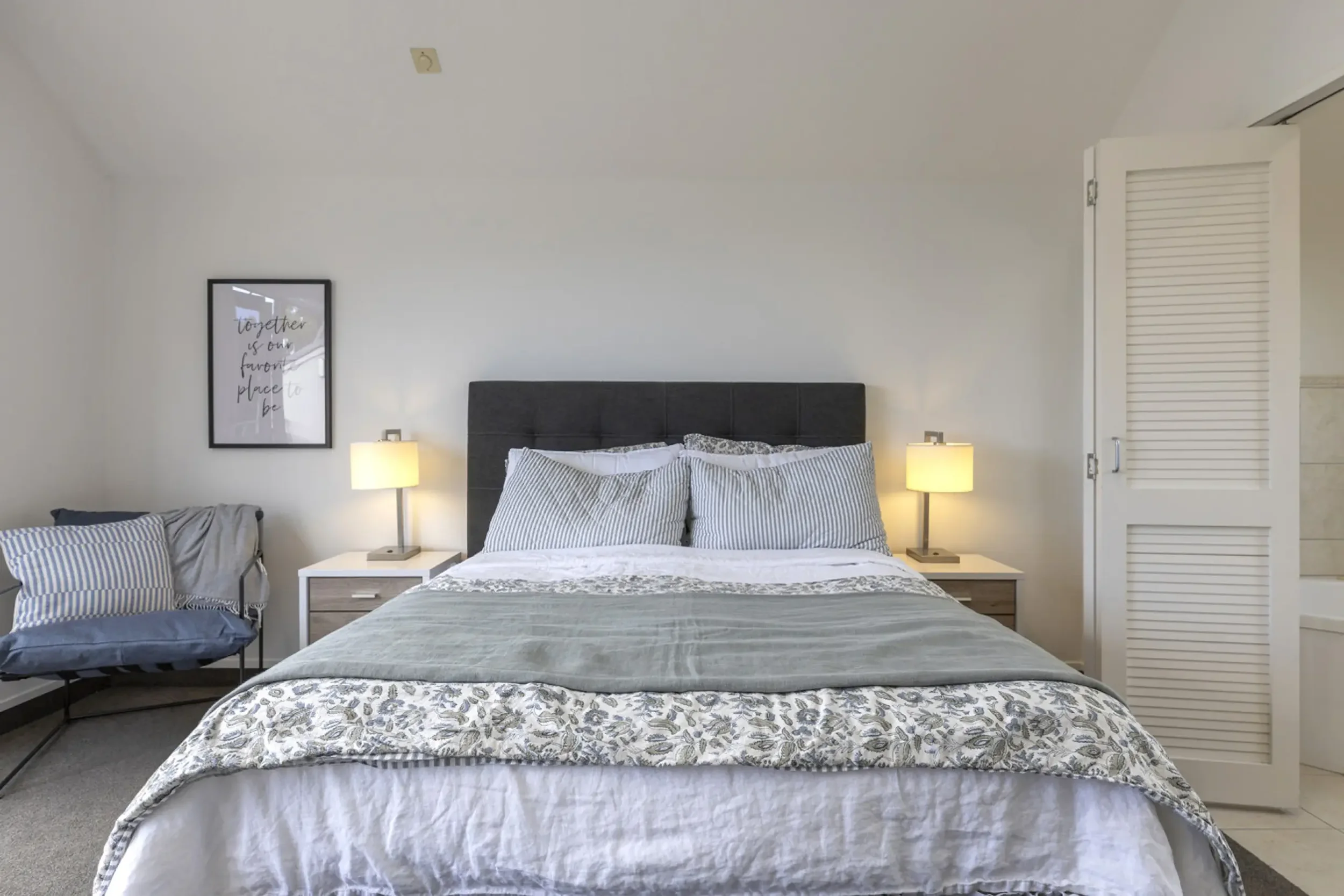 A neatly made bed with a dark gray upholstered headboard, two bedside tables with lamps, two striped pillows, and a white framed wall art. There is a chair with a matching striped pillow and a gray throw blanket on the left side of the bed, and a whi