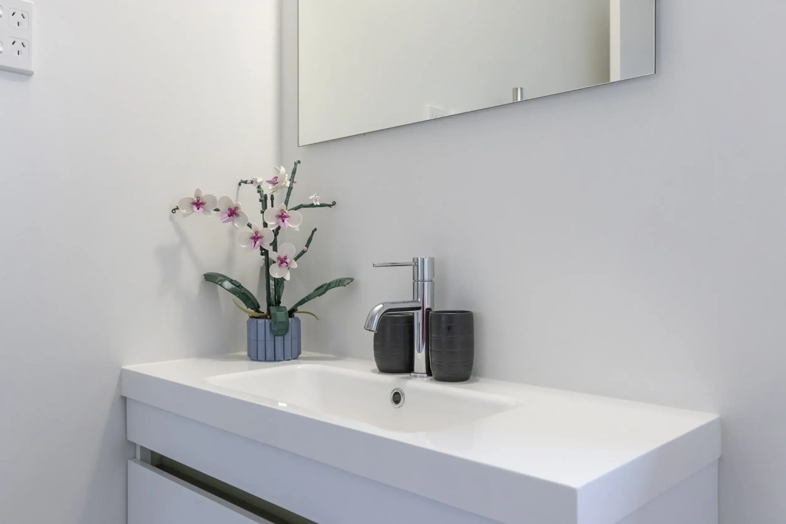 Modern bathroom sink with a mirror, a blue vase with artificial pink and white orchids, and two black cups.