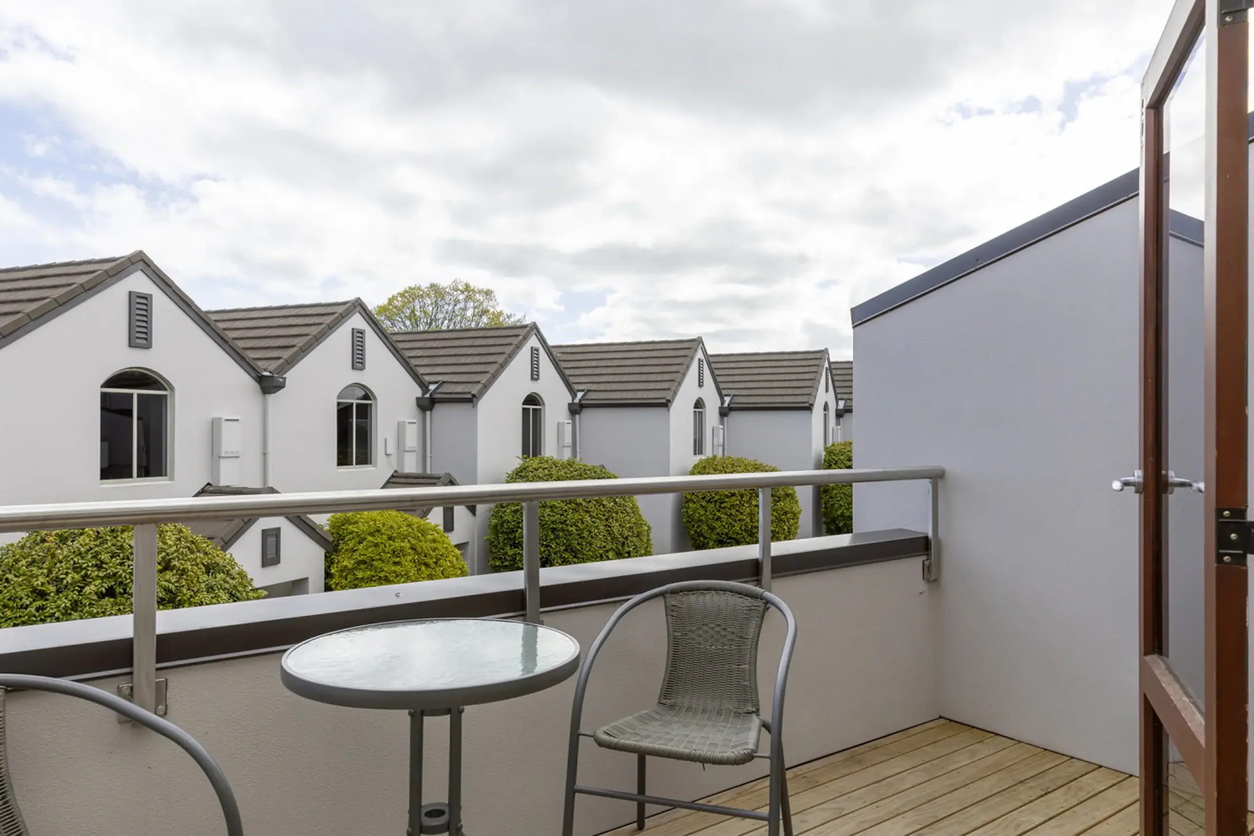 View from a balcony featuring a small round glass table and a metal chair with tan woven seat, overlooking a row of modern white townhouse buildings with gray roofs, green bushes, and a partly cloudy sky.