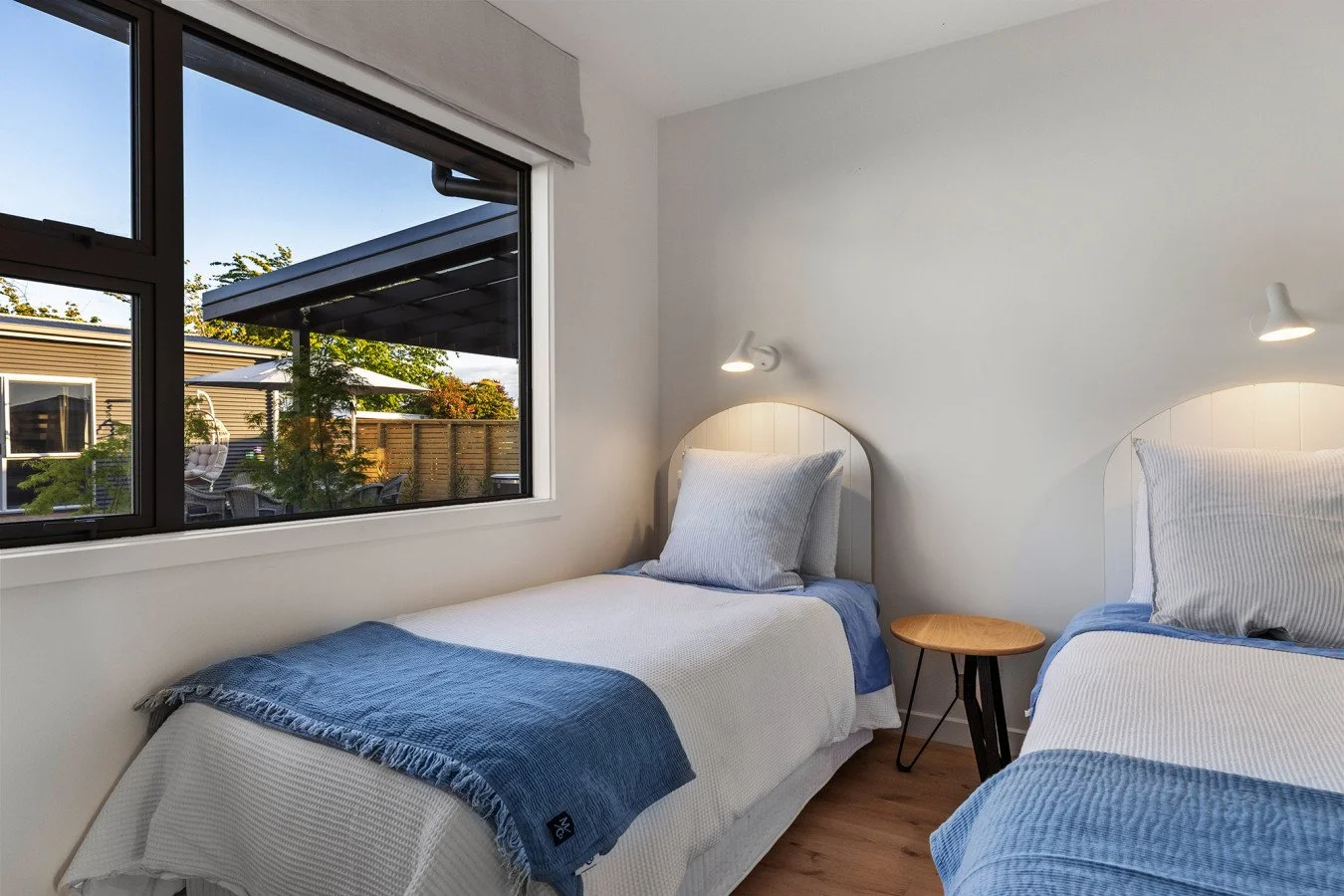 A bedroom with two single beds with white bedding and blue throws, a small round wooden table between the beds, a window showing an outdoor patio with seating and greenery, and wall-mounted reading lights above each bed.