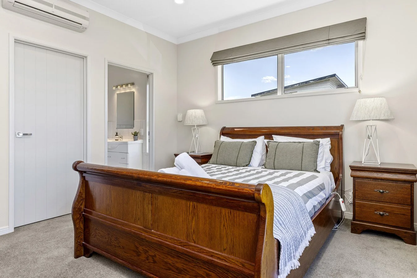 Bedroom with wooden bed frame, gray pillows, white bedding, bedside table lamps, window with blinds, and an ensuite bathroom visible through an open door.