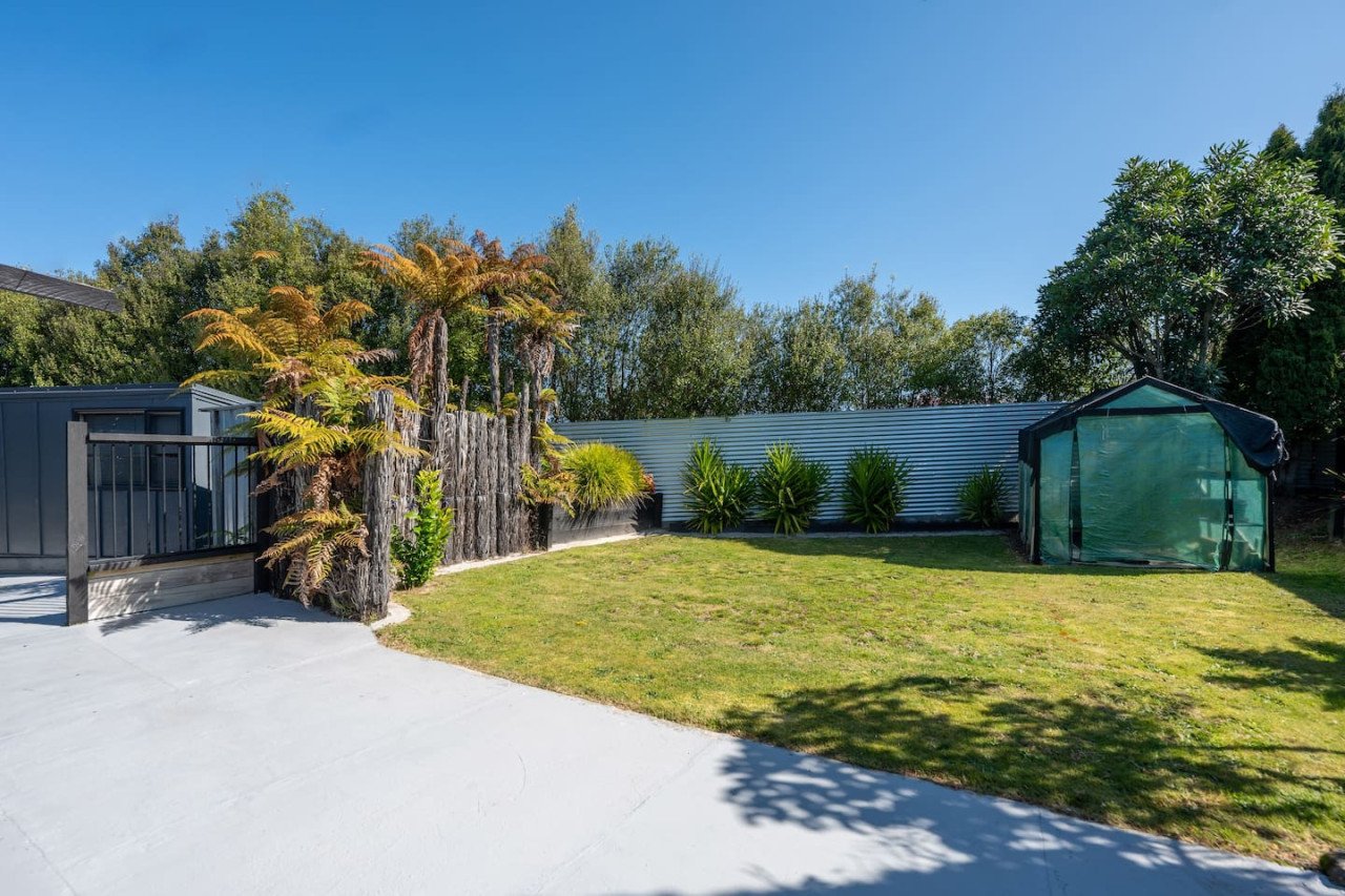 A backyard with a small green greenhouse, a wooden fence, and green plants against a metal fence, under a clear blue sky.