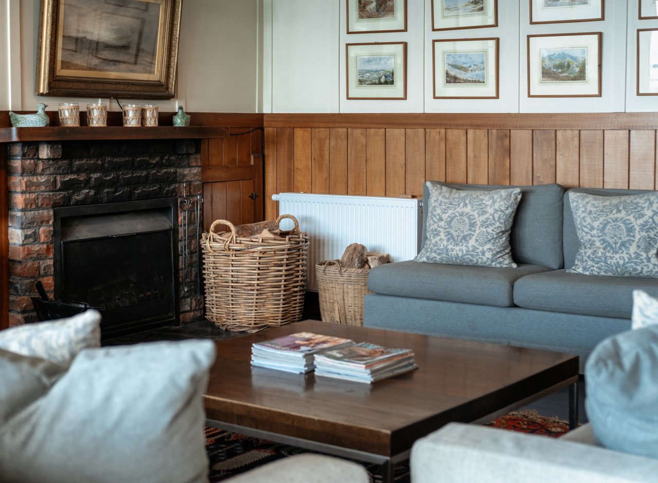 Living room with a brick fireplace, framed artwork on the walls, a grey couch with patterned cushions, a wooden coffee table with magazines, and wicker baskets filled with firewood.