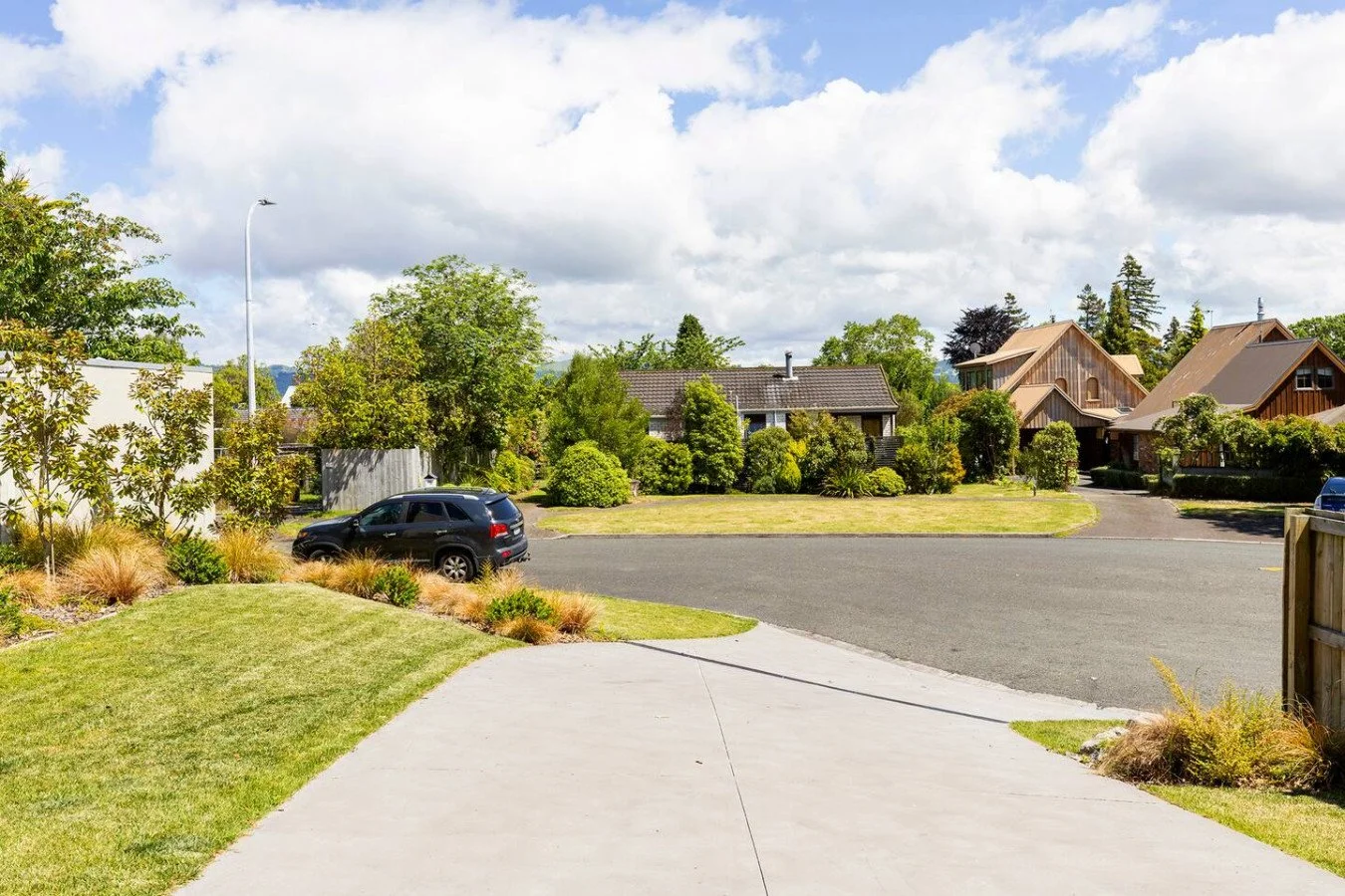 Residential neighborhood with a paved driveway, parked cars, green lawns, trees, and houses under a partly cloudy sky.
