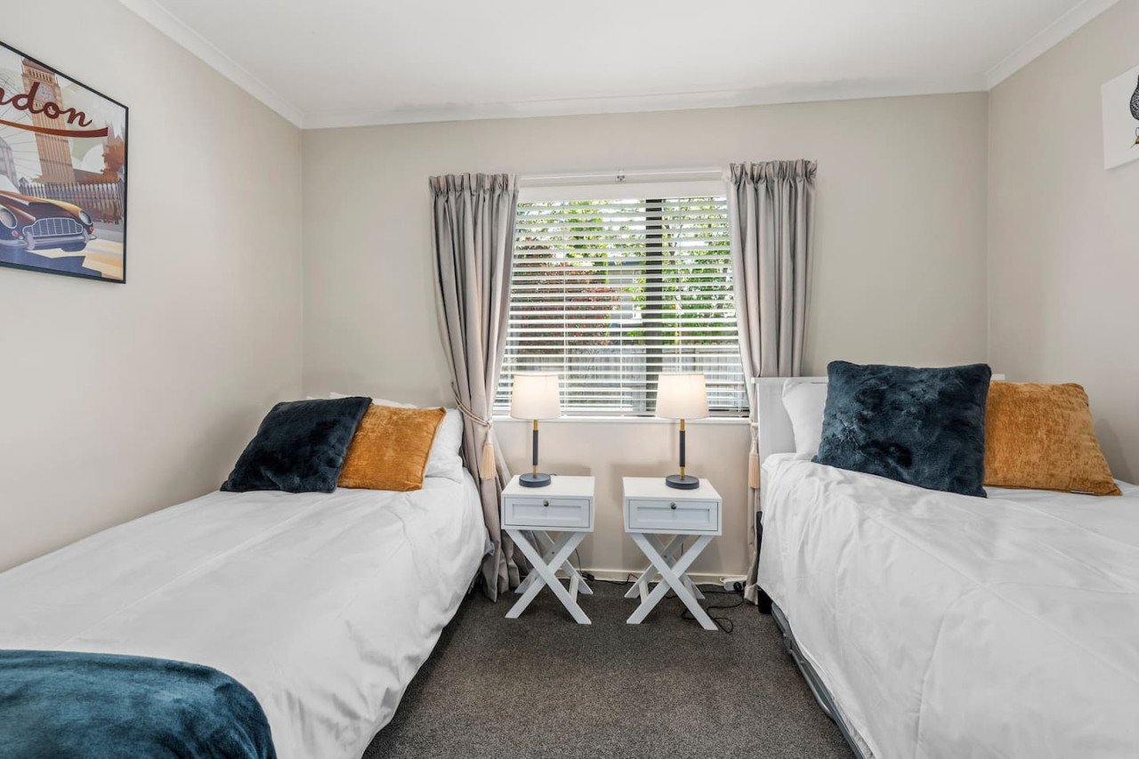 A bedroom with two single beds, each with dark blue and orange pillows, a window with blinds and curtains, and two small white nightstands with lamps between the beds.