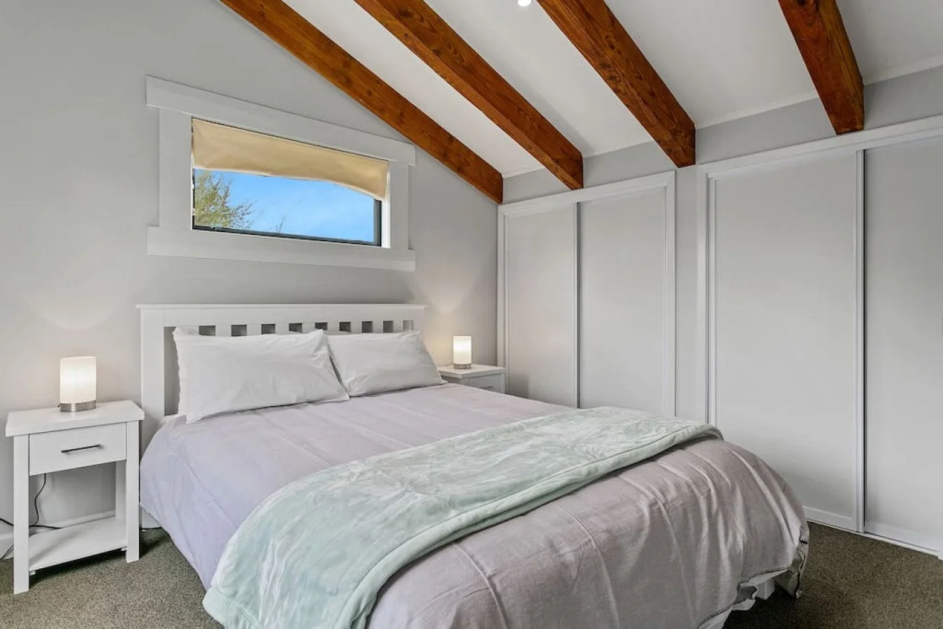 A bedroom with a white bed, two white nightstands with lamps, a window with a beige shade, white walls, and exposed wooden ceiling beams.