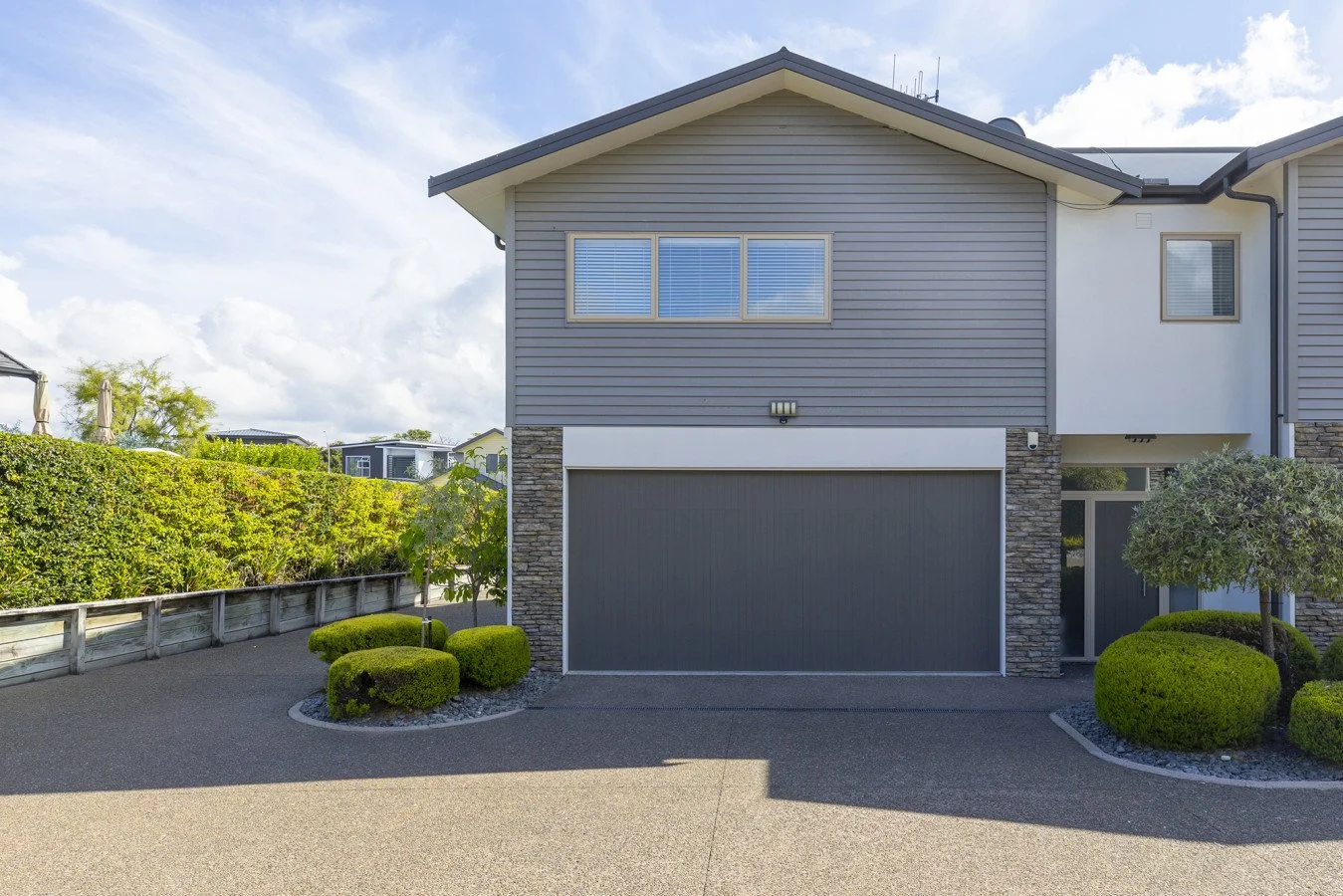 A modern two-story house with gray siding, stone accents, and a garage door. Well-maintained landscaping with shrubs and trees in the front yard.