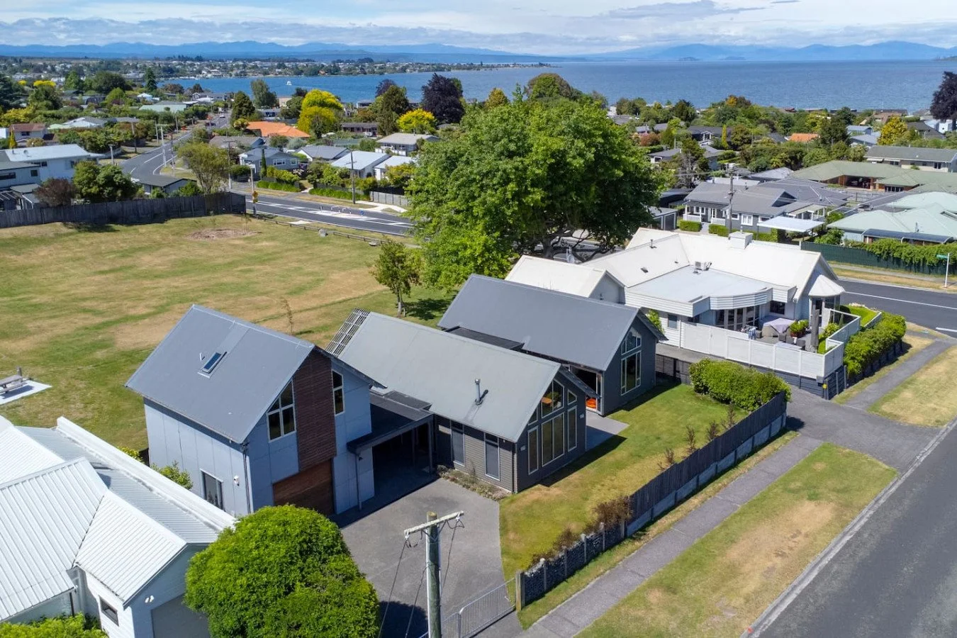 Aerial view of a residential neighborhood near a body of water, showing houses with metal roofs, a large grassy area, trees, streets, and a lake in the background.