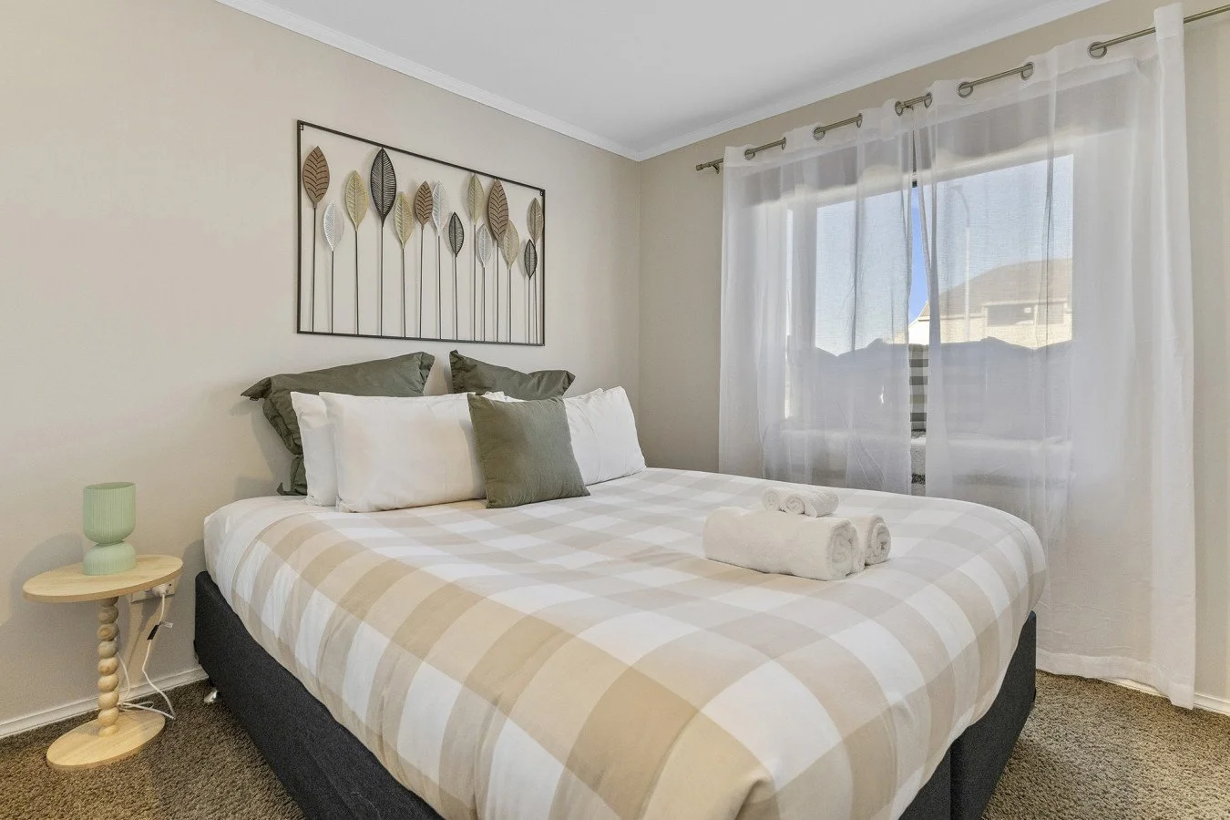 A neatly made bed with white and beige checkered bedding, dark gray pillows, and white towels with a bedroom window with sheer curtains.