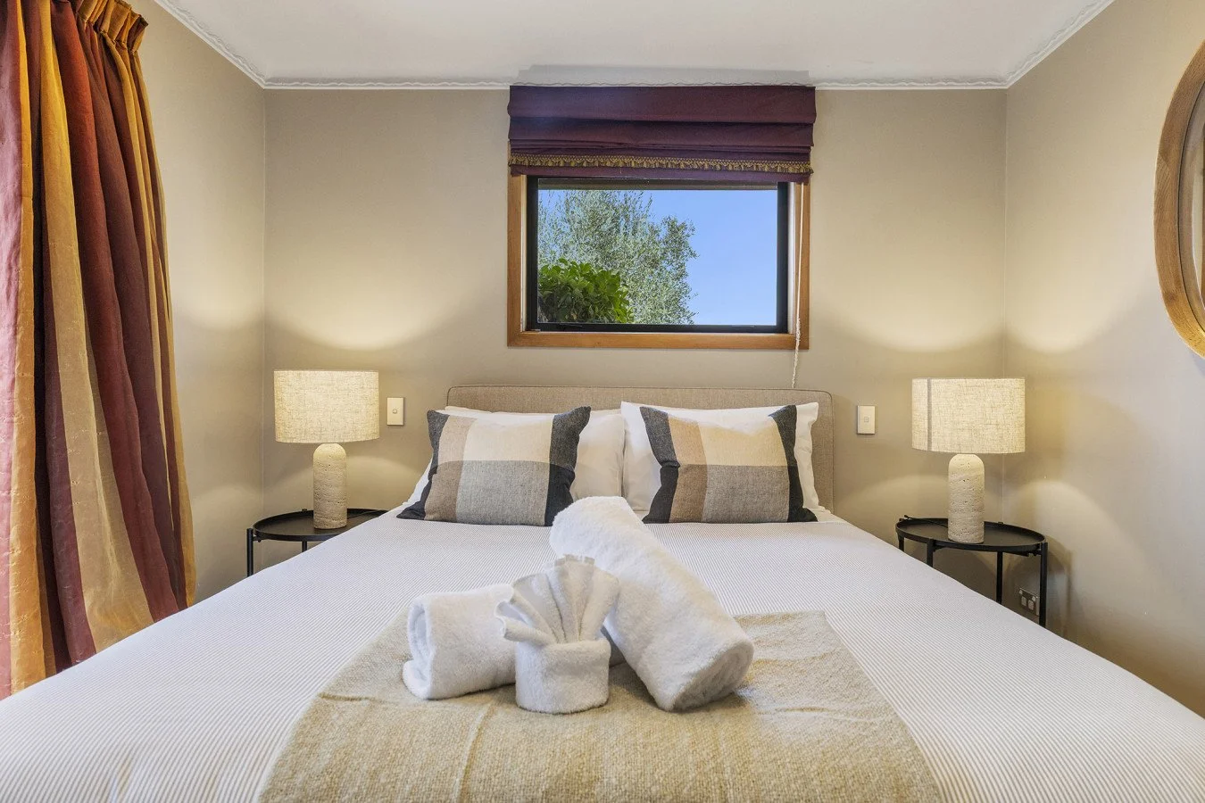 A neatly made bed with white bedding, two pillows with checkered covers, and folded towels shaped like a flower on a beige blanket, in a bedroom with beige walls, bedside lamps, a small window showing trees outside, and drapes on the sides.