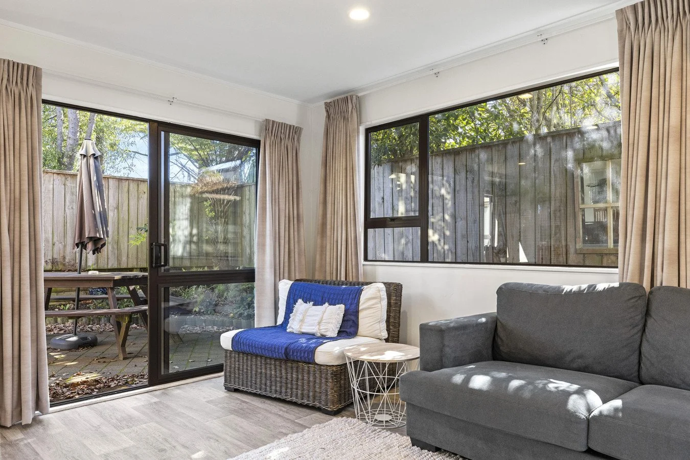 Living room with gray sofa, wicker chair with blue throw and pillows, large windows with beige curtains, sliding glass door leading to outdoor patio with table, umbrella, and wooden fence, sunlight streaming in.