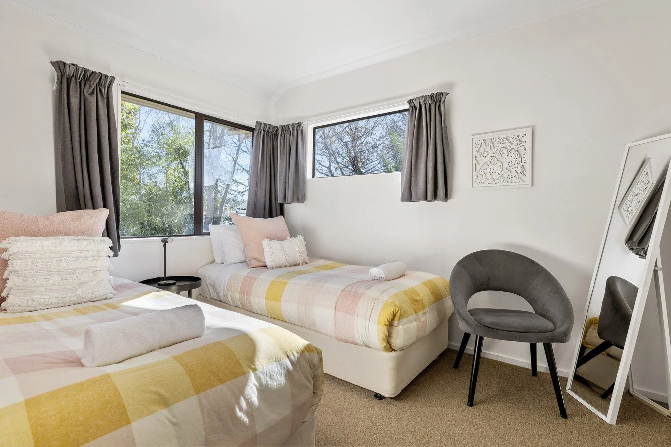 A bright bedroom with two single beds that have pastel plaid bedspreads, pillows, and rolled towels. There are large windows with gray curtains, a small black bedside table, a gray chair, wall art, a full-length mirror, and a view of trees outside.