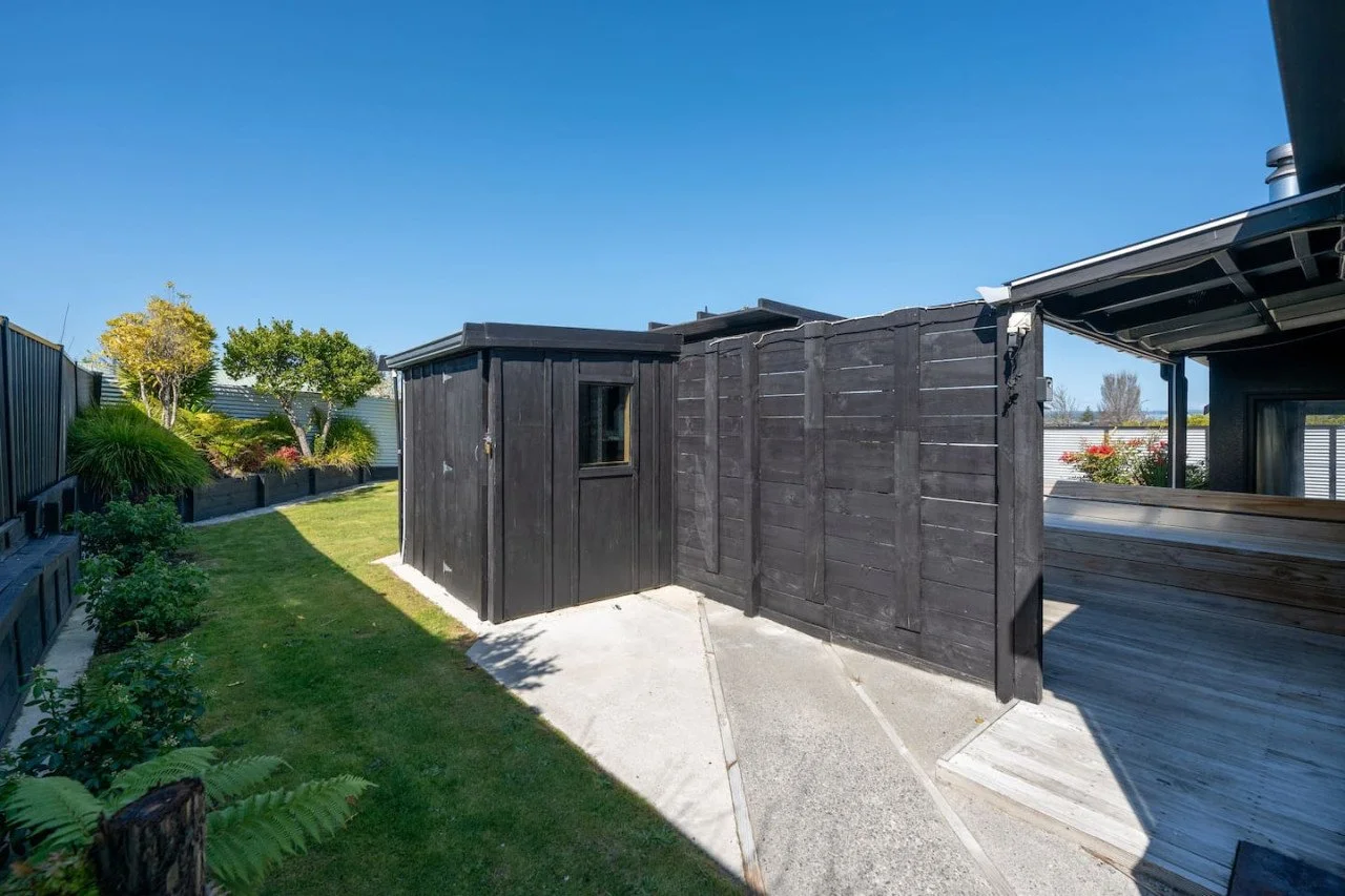 A backyard with a small black shed, a black wooden privacy fence, green lawn, plants, bushes, trees, and a wooden deck in bright sunlight under a clear blue sky.