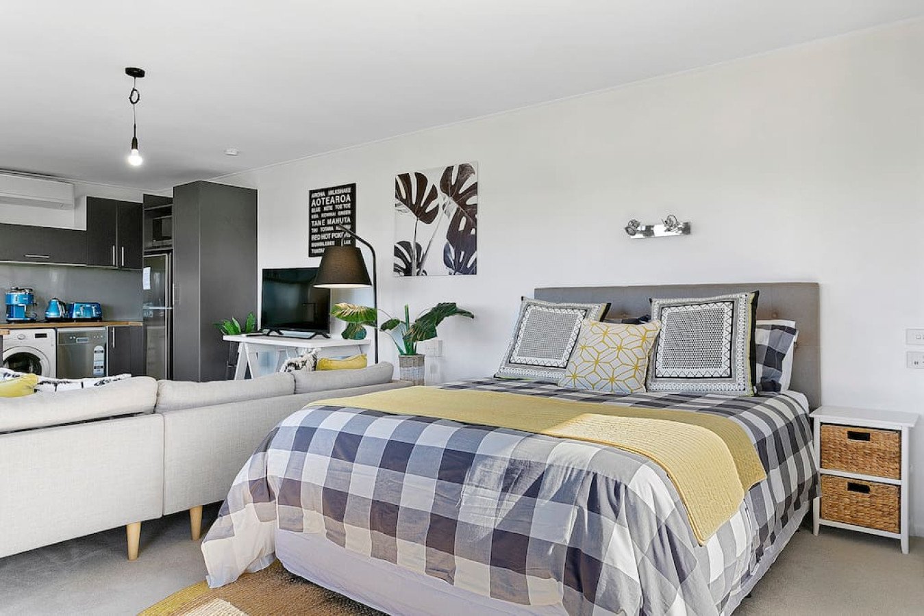 Modern studio apartment with a bed, white nightstand with woven baskets, a TV on a white table, plants, black and white wall art, and a small kitchen area with blue appliances.
