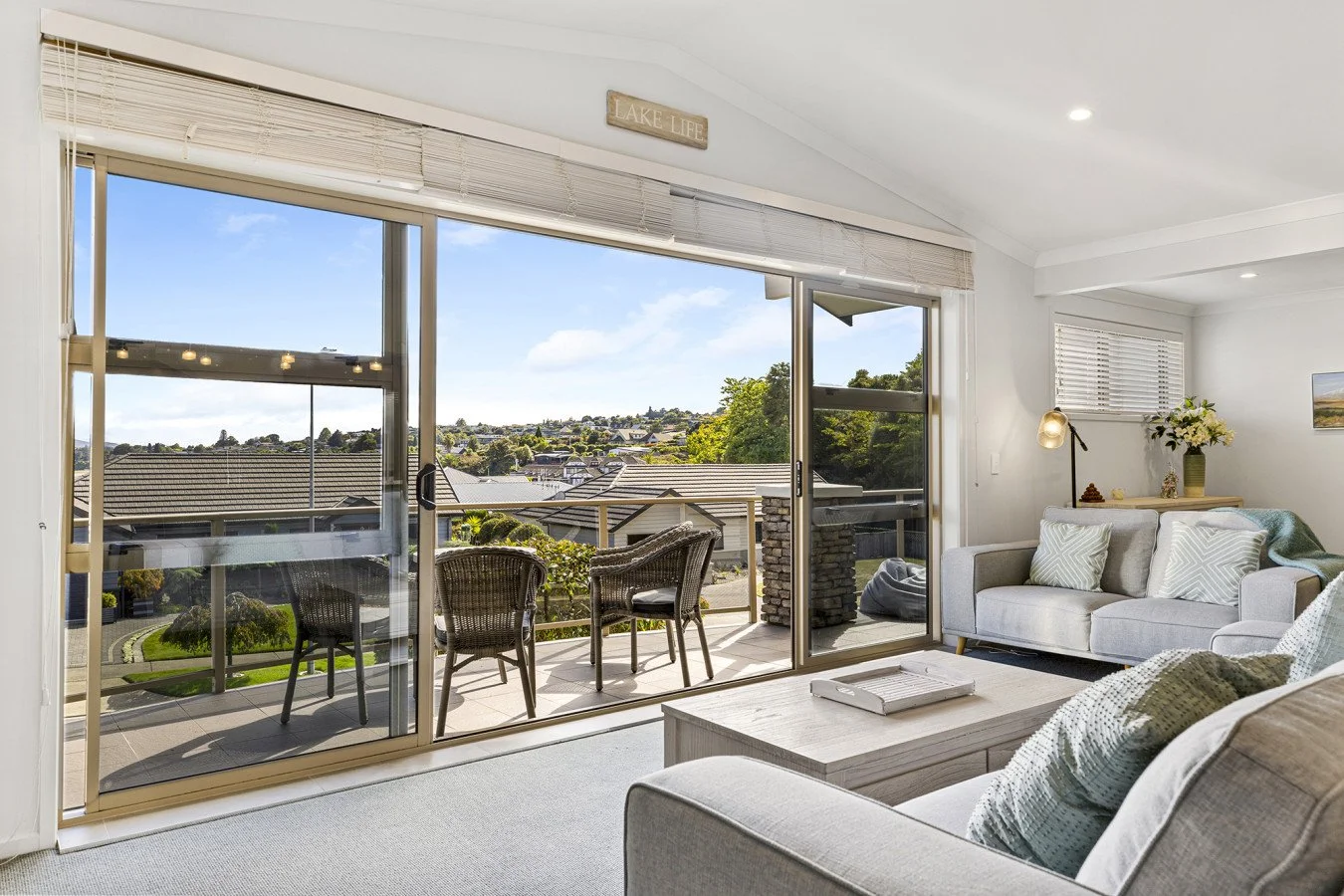 Living room with sliding glass doors leading to a balcony overlooking a neighborhood with hilly terrain, white walls, white couches, and a wooden coffee table.