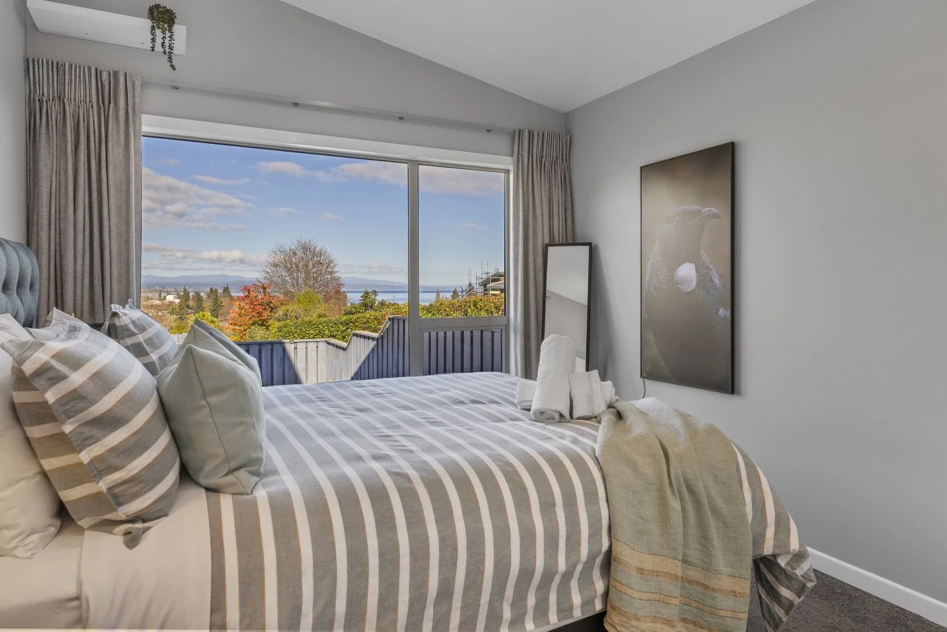 Bedroom with large window showing outdoor view, neatly made bed with striped and plain pillows, beige and gray bedding, wall art, and a standing mirror.