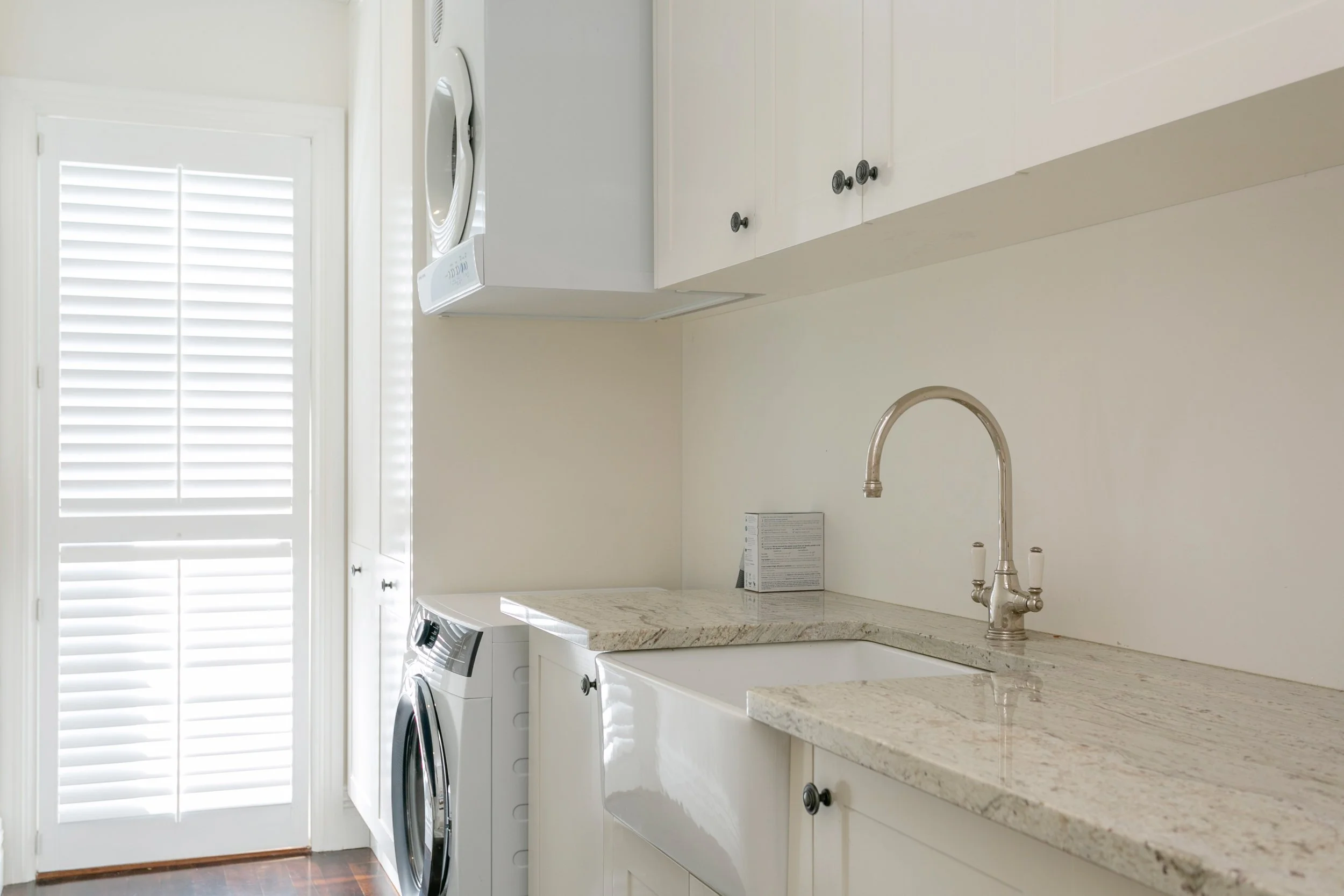 Laundry room with white cabinets, a granite countertop, a large white farmhouse sink, a silver faucet, a washing machine, a dryer, and a door with blinds.