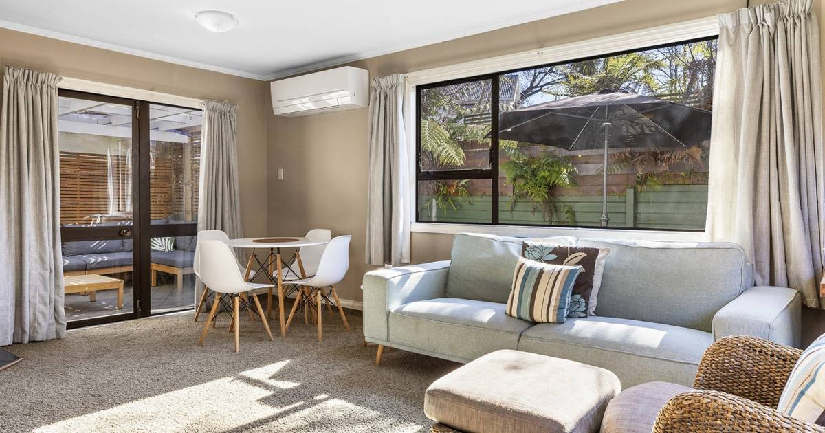 Living room with a light gray sofa, striped and floral pillows, a beige ottoman, a woven chair, large windows with curtains, a dining table with four white chairs, and a sliding glass door leading outside to a patio with an umbrella and greenery.