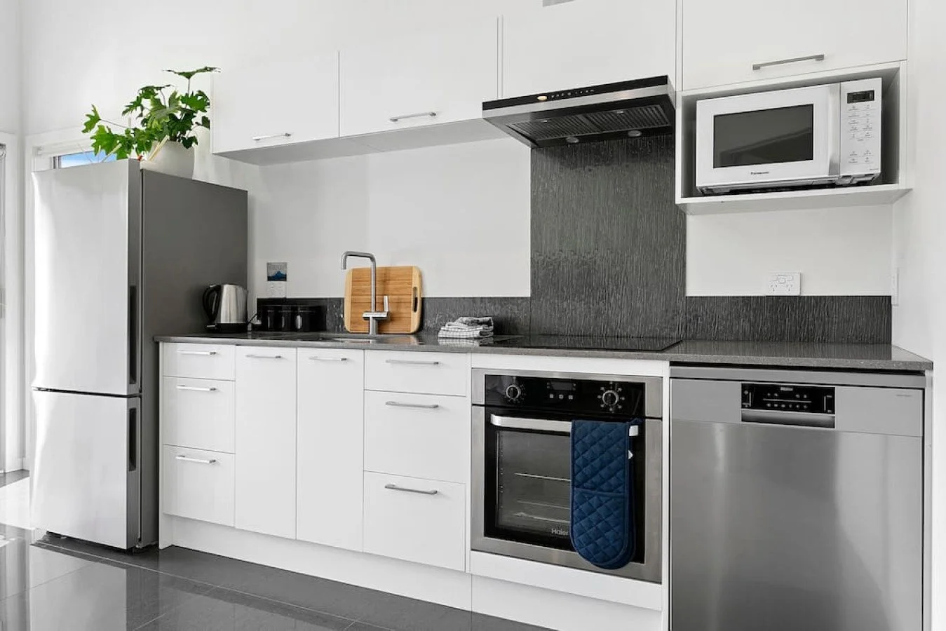 Modern kitchen with white cabinets, black countertops, a stainless steel refrigerator, microwave, oven, and dishwasher, with a potted plant on top of the refrigerator.