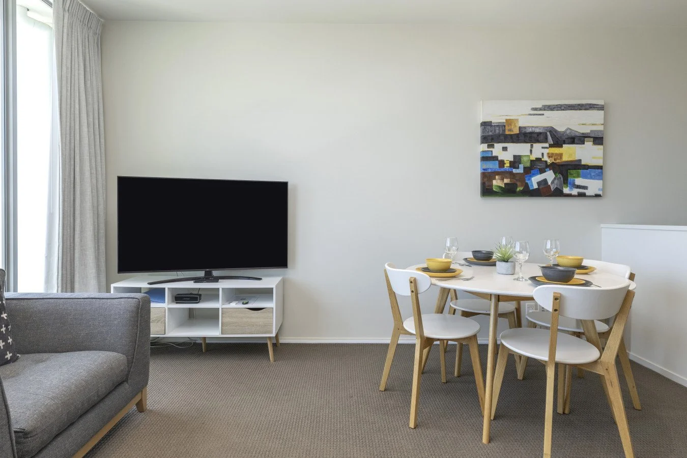 Living room with a gray couch, a white TV stand with a flat-screen television, a round dining table with four white chairs, and a painting on the wall.