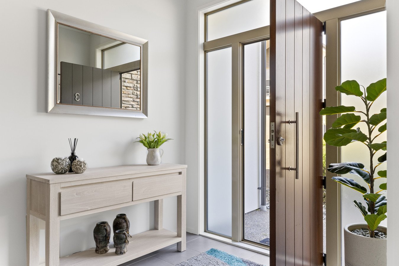 Entryway with a door, a mirror on white wall, a white console table with vases and a potted plant, and a large potted plant in a corner.