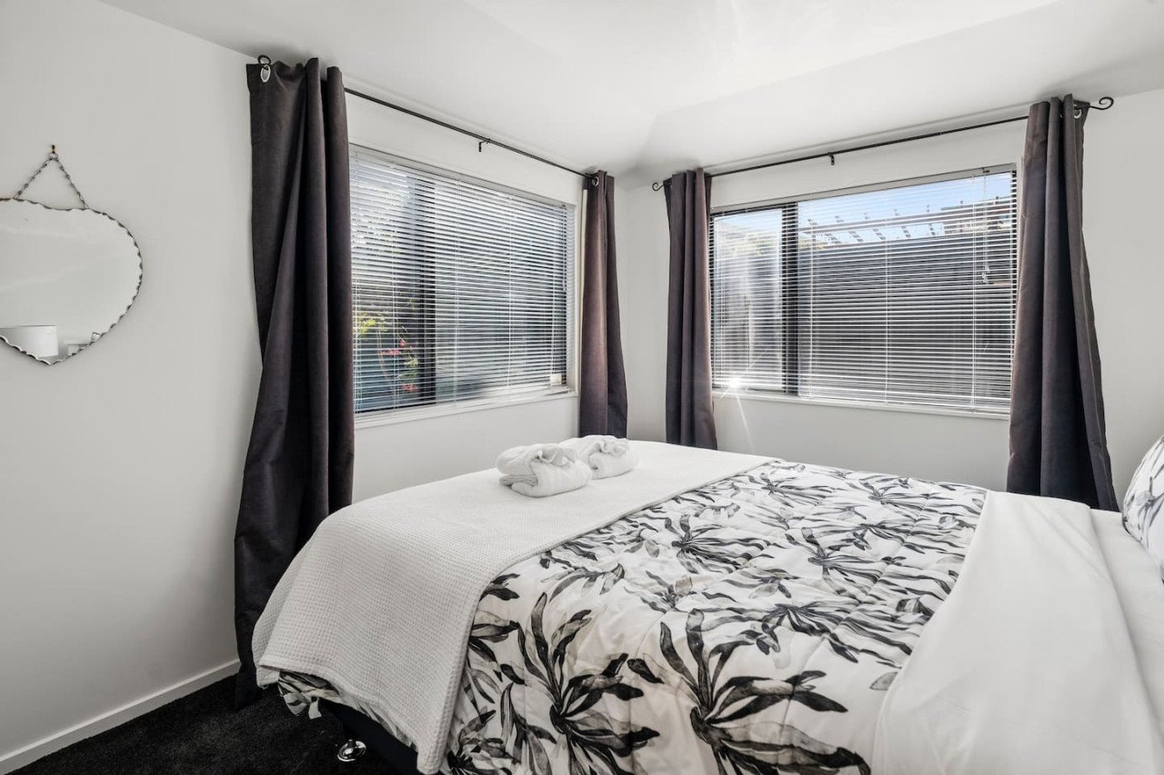 A neatly made bed with white and gray floral bedding, two towel swans on top, and two large windows with blinds and gray curtains in a bright, clean bedroom.
