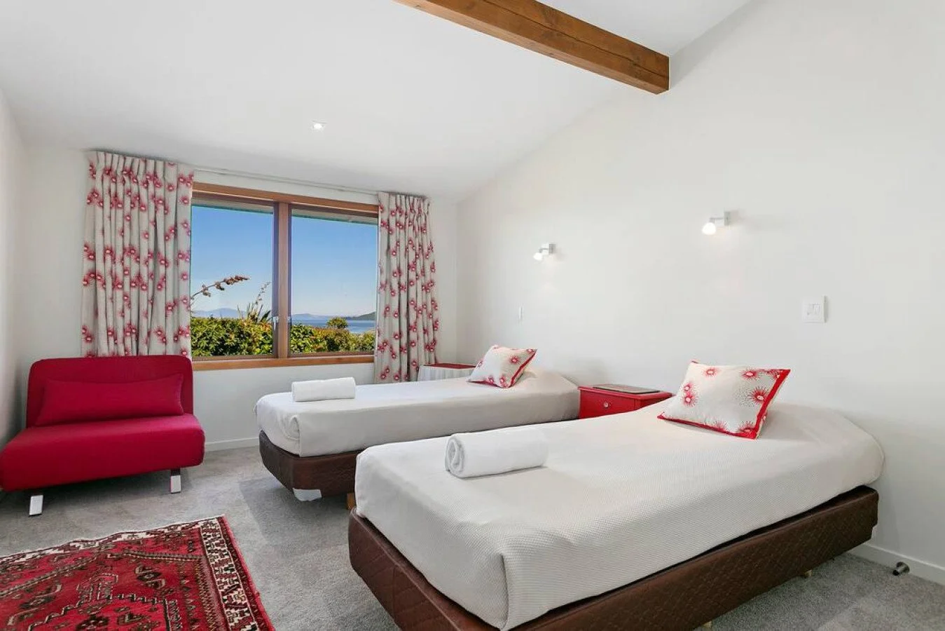 A bright hotel room with two beds dressed in white sheets and pillows with red floral pillowcases. There is a red sofa in the corner, a window with floral curtains, and a view of greenery and blue sky outside.