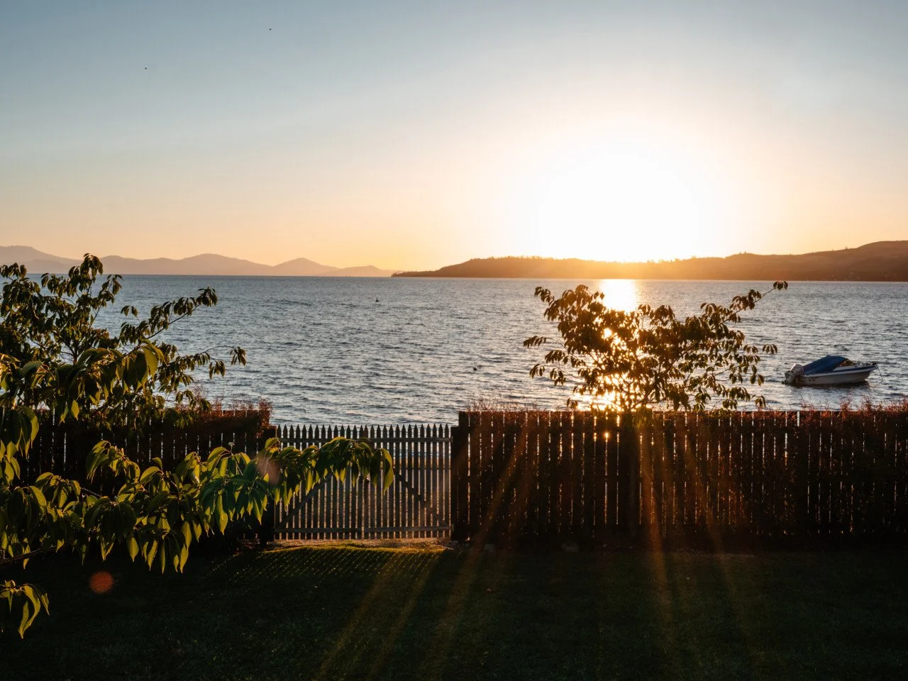 Sunset over a lake with a boat near the shore, trees, and a wooden fence in the foreground.